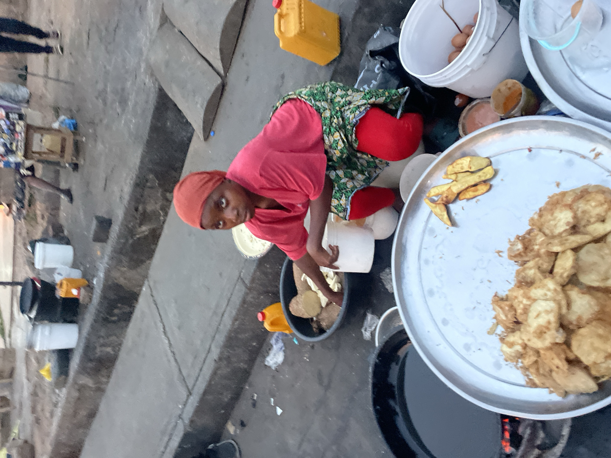 A person in Jos, Nigeria is kneeling on the ground. They are wearing a red shirt, red leggings, and a green and white floral skirt. They have a red head wrap. They are reaching into a grey bin with their left hand, which appears to contain sliced sweet potatoes. A small yellow jerrycan is adjacent to the bin. In front of the person is a large, round, silver tray filled with fried sweet potatoes, along with a smaller portion of fried potatoes on the side. To the right is a large metal pot. Other items nearby include a white bucket filled with eggs, a black bag, and other containers. The ground is grey and appears to be concrete.
