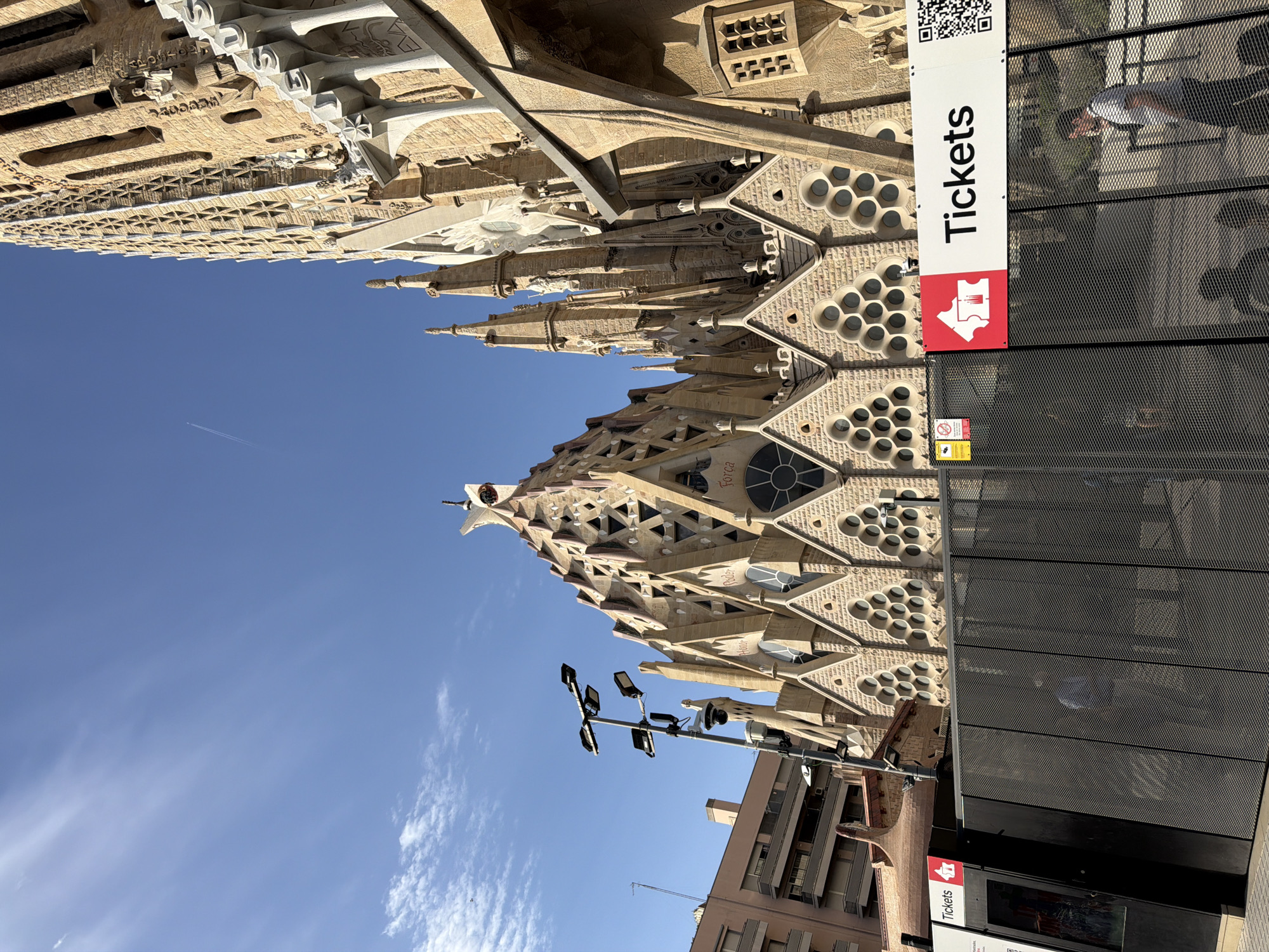 The image shows a side view of the Sagrada Familia basilica in Barcelona, Spain, under a bright blue sky with scattered clouds. The basilica's distinctive, organic architecture is visible, characterized by intricate stonework and pointed spires. In the foreground, a black metal fence runs across the bottom of the image, partially obscuring the view of the basilica's base. Above the fence, a white banner with red accents reads "Tickets" and features a ticket icon. To the left of the banner is a black booth with the word "Tickets" displayed on it. Several people are visible in the background, appearing to be pedestrians or tourists. One person in a light-colored shirt walks away from the camera on the right side of the image. The overall scene suggests a day of tourism or activity outside the famous landmark.