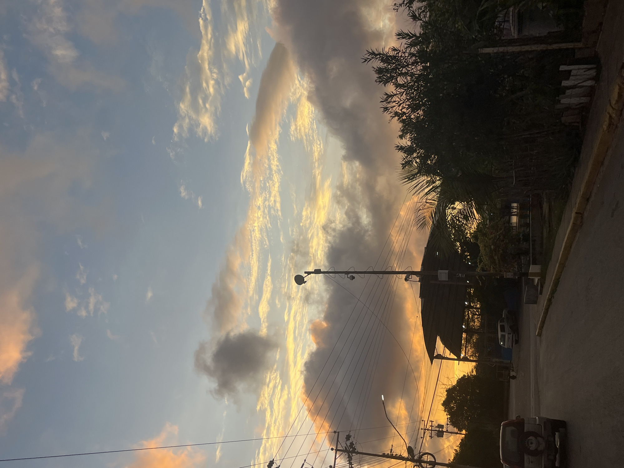 The image captures a serene sunset scene in Santa Ana, Venezuela. The sky is a canvas of warm oranges, pinks, and blues, with clouds casting dramatic shadows. In the foreground, a paved road leads into the distance, flanked by a sidewalk and lush vegetation.
On the left side of the road, a silver SUV is parked. Further down the street, a covered structure with a dark roof is visible, and a white pickup truck is parked nearby. Utility poles with numerous wires stretch across the sky, adding a touch of urban life to the natural beauty of the sunset.
The overall impression is one of peace and tranquility, a typical evening in a Venezuelan town.