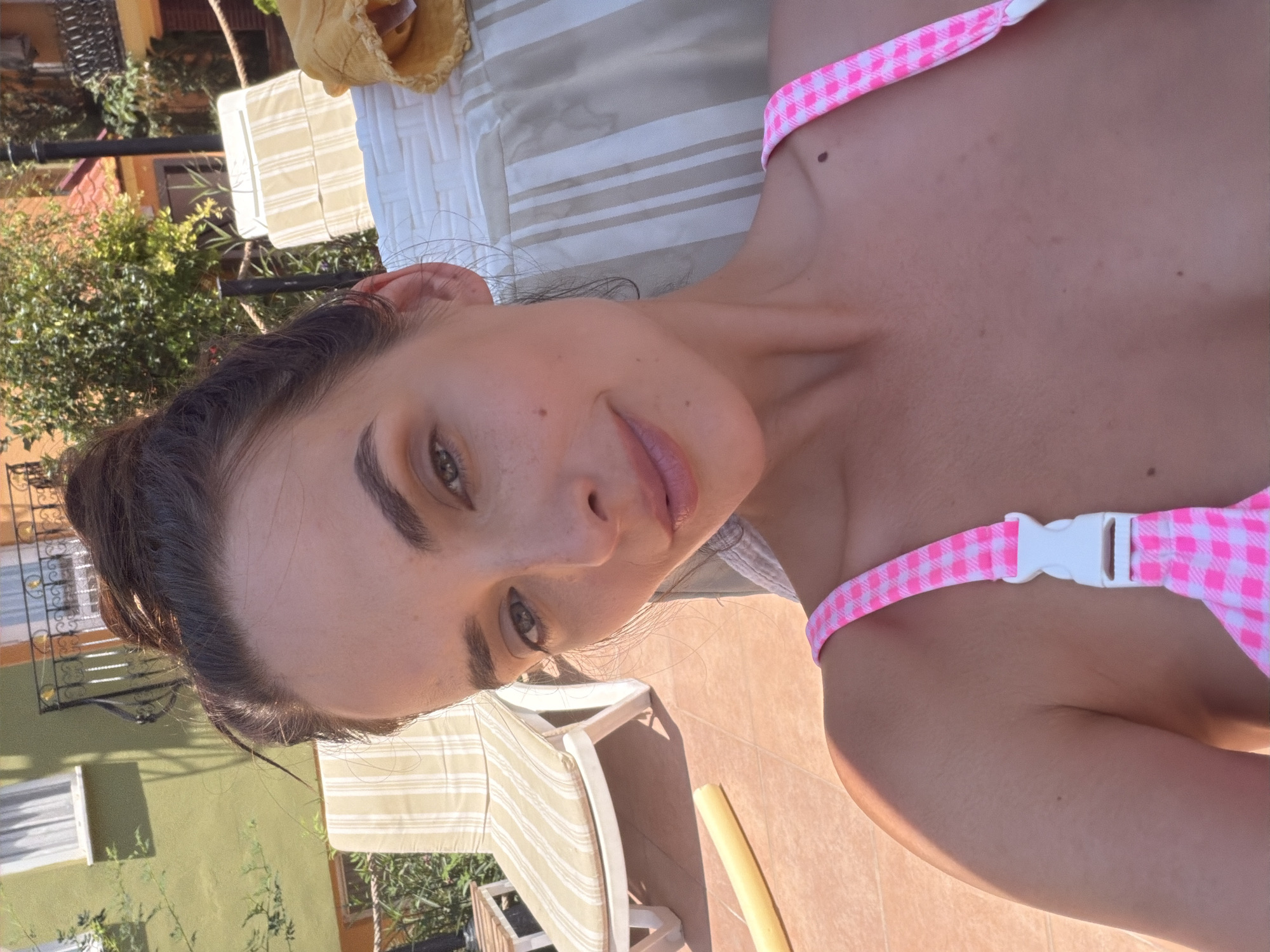 In Fethiye, Turkey, a young woman with her hair in a bun is seen in a close-up selfie, wearing a pink and white checkered bikini top. She is seated outdoors, with sun loungers and a building visible in the background, suggesting a poolside or beach resort setting. The lighting indicates it is daytime.