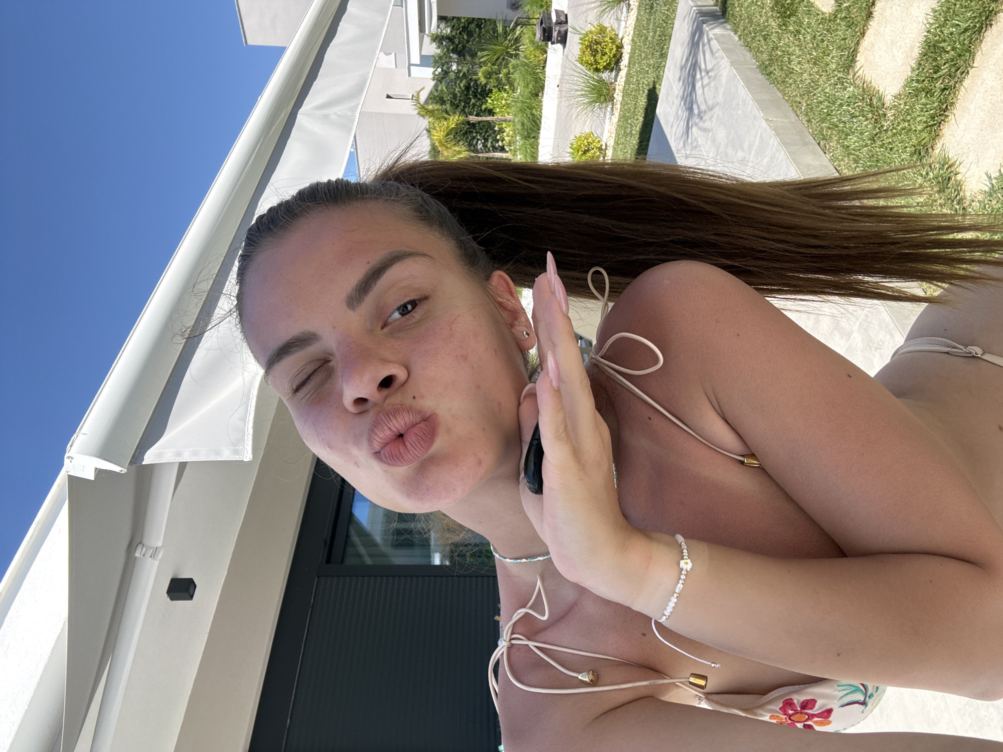 A young, light-skinned woman is featured in a selfie-style photo taken outdoors. She has brown hair tied in a high ponytail, is winking, and puckering her lips as if giving a kiss. Her left hand is raised to the side of her face, holding a black object. She is wearing what appears to be a light-colored floral bikini and is adorned with a white beaded bracelet and necklace.
The background includes a white building with black-framed windows, green grass, and stone pathways. The location appears to be a sunny outdoor area, possibly a garden or poolside. The sky is blue.