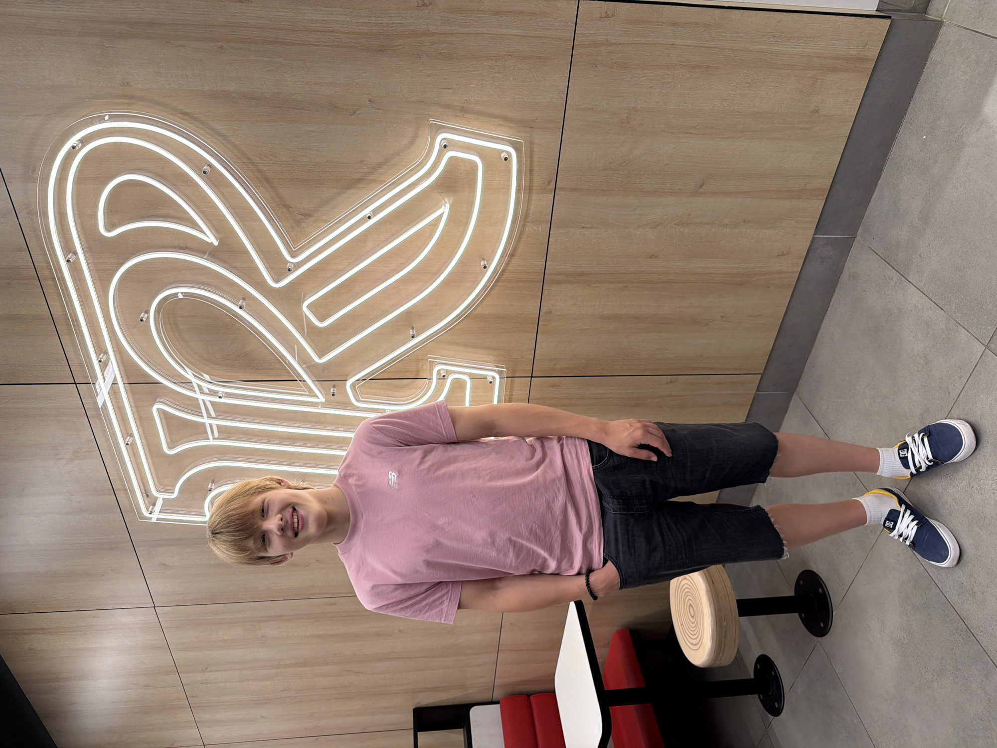 A young man with blonde hair stands in front of a large neon sign shaped like the letter "R" in a restaurant. He is wearing a pink t-shirt, dark denim shorts, white socks with yellow accents, and blue and white sneakers. He has his hands in his pockets and is smiling at the camera. To his left, there is a booth with red seating and a white table. Further to the left, a round stool with a wooden top is visible. The wall behind the man is paneled with light wood and the floor is tiled in gray. The neon sign is white and outlined with a clear plastic casing. The restaurant appears to be a casual dining establishment. The specific location within Izhevsk, Russia, cannot be determined from the image alone.