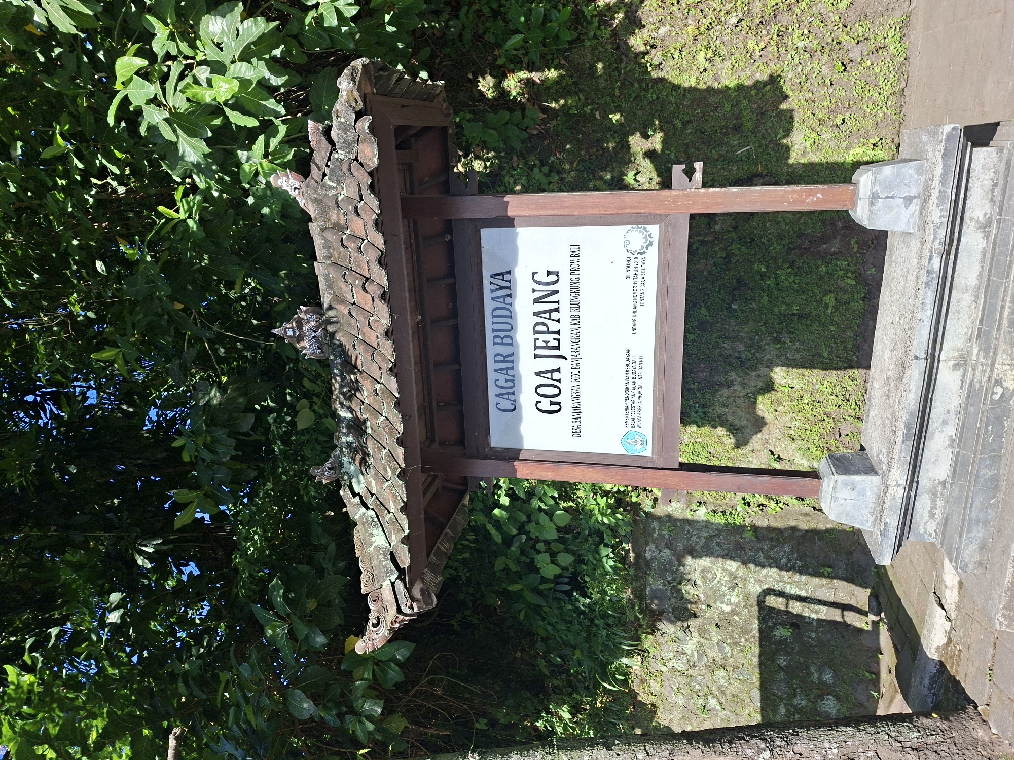 In Banjarangkan, Indonesia, a cultural heritage site sign stands under a traditional tiled roof, supported by wooden posts. The sign, primarily white with dark text, reads "CAGAR BUDAYA GOA JEPANG" and further specifies "DESA BANJARANGKAN, KEC. BANJARANGKAN, KAB. KLUNGKUNG, PROV. BALI". Below this, smaller text details government ministries and regulations. The structure is situated outdoors, with lush green trees and foliage forming a dense backdrop. To the left, a portion of a tree trunk is visible. The ground in front of the sign is paved with stone tiles, and a textured stone wall covered in moss and small plants is behind it. Sunlight casts shadows, indicating a bright, clear day. The scene is static, with no people or discernible activity.