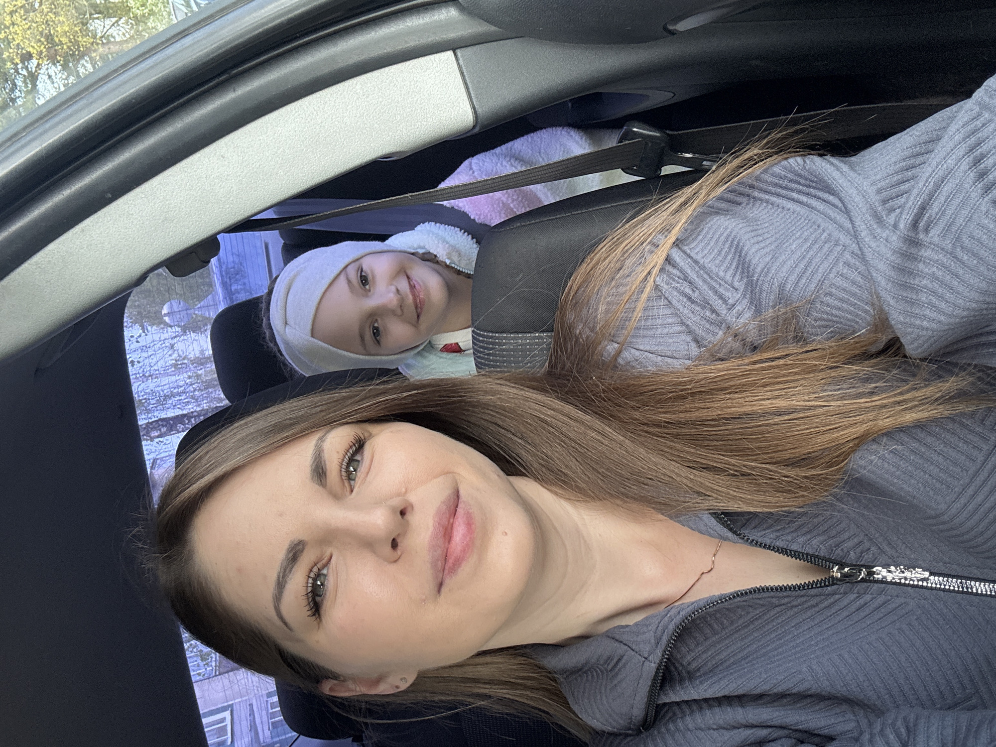 Two individuals are visible inside a vehicle. One is an adult female, positioned in the foreground, wearing a gray zip-up jacket and has light brown hair. The other is a child, seated in the back, wearing a white head covering and a colorful jacket, secured with a seatbelt. The interior of the vehicle is predominantly gray. The background, seen through the windows, suggests an urban setting. Based on the location metadata, this scene is situated in Раздолье, Russia.