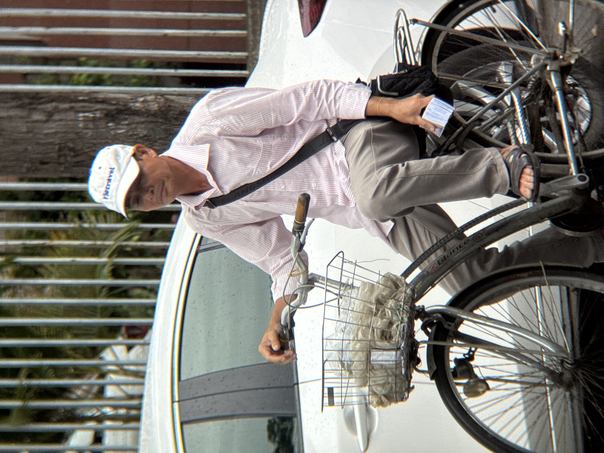 A man wears a white cap with the word "fleetravel" and a pink and white striped shirt as he sits on a bicycle. The bicycle's front basket is filled with plastic bags. The man is holding a stack of cards in his right hand. He is wearing grey pants and black sandals. Behind him, a white car is parked. The scene appears to be on a street in Nha Trang, Vietnam, with a metal fence and trees visible in the background.