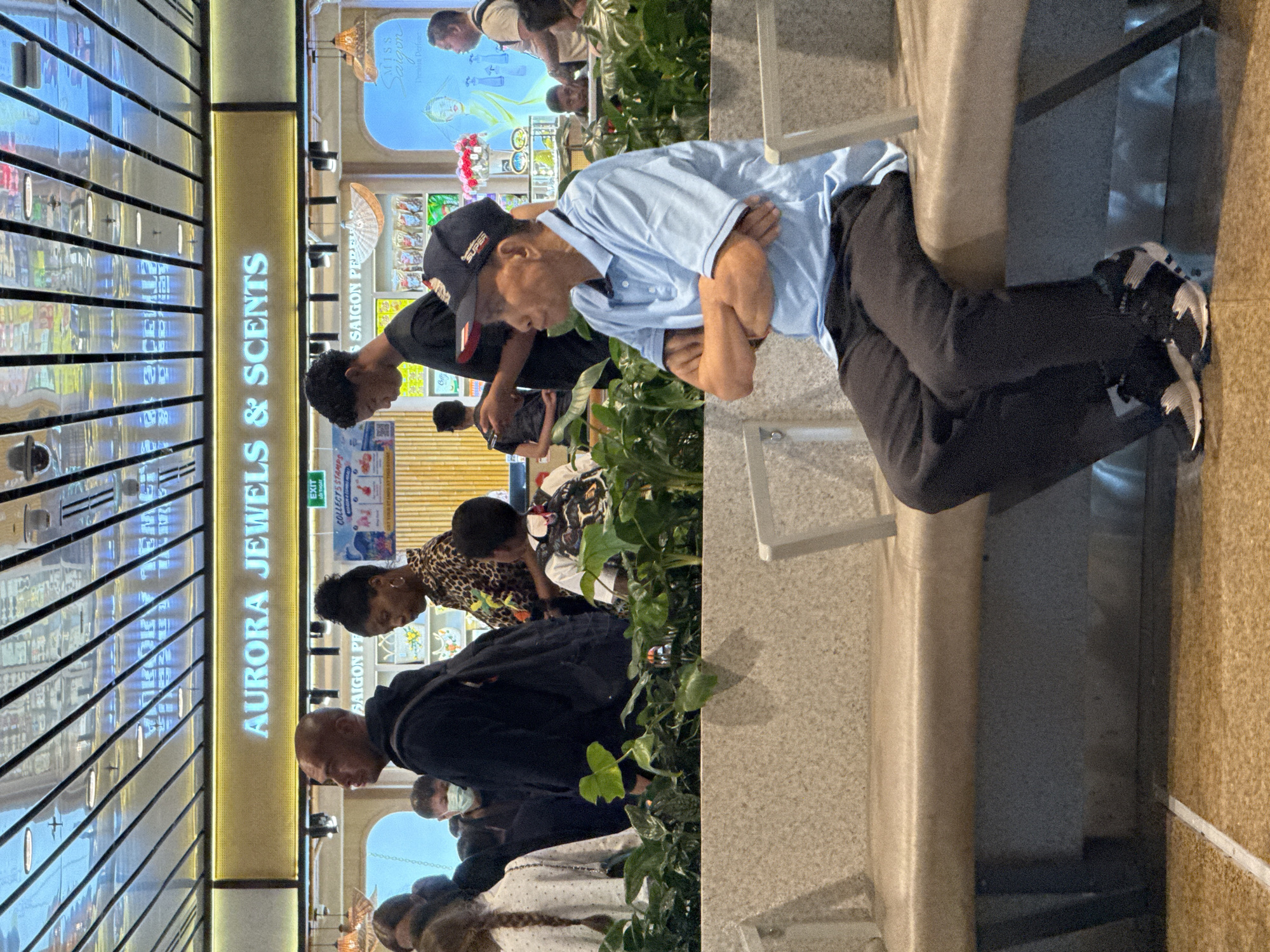 In Bắc Cam Ranh, Vietnam, a group of people are seen in an indoor commercial space, likely an airport or shopping mall, as indicated by the "AURORA JEWELS & SCENTS" signage and the reflective ceiling panels. In the foreground, an elderly man with dark skin and thinning grey hair, wearing a light blue polo shirt, dark pants, and a black baseball cap with red trim and "SUPER" text, is seated with his arms crossed. He is looking to his left. Behind him, a variety of individuals are visible, including a woman with dark, curly hair wearing a leopard print top, a person with a medical mask, and a young person with a backpack. The environment includes a low, stone bench, lush green plants, and displays of products and advertisements in the background. The lighting suggests an indoor setting with overhead illumination and some decorative lamps.