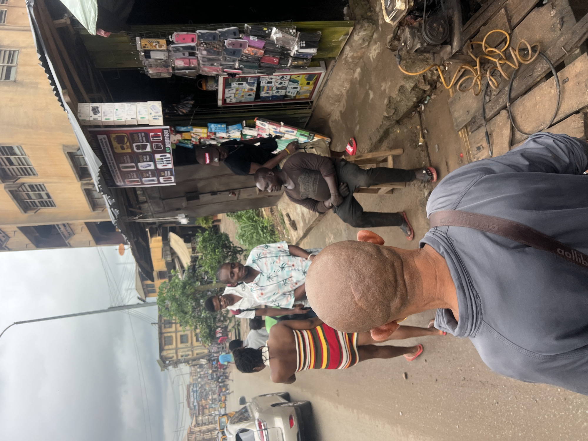 The image depicts a street scene in Nkpor, Nigeria, on a cloudy day. In the foreground, a person's back is visible, wearing a gray shirt with a brown strap across their shoulder. They are facing a street where several individuals are present.
To the left of the foreground figure, a woman in a striped dress walks away from the camera. Behind her, a man wearing a patterned short-sleeved shirt and another man in a white shirt are walking. Further down the street, various vehicles and buildings can be seen, suggesting a busy urban environment.
To the right of the street, a vendor stall displays numerous cell phone accessories, including cases and chargers. Two men are present at the stall: one is seated on a wooden stool, wearing a dark long-sleeved shirt and black pants, while the other stands behind him, wearing a black t-shirt and a red baseball cap. A generator and various tools are visible on the ground near the stall. The architecture of the buildings in the background is typical of the region, with multiple stories and balconies.
