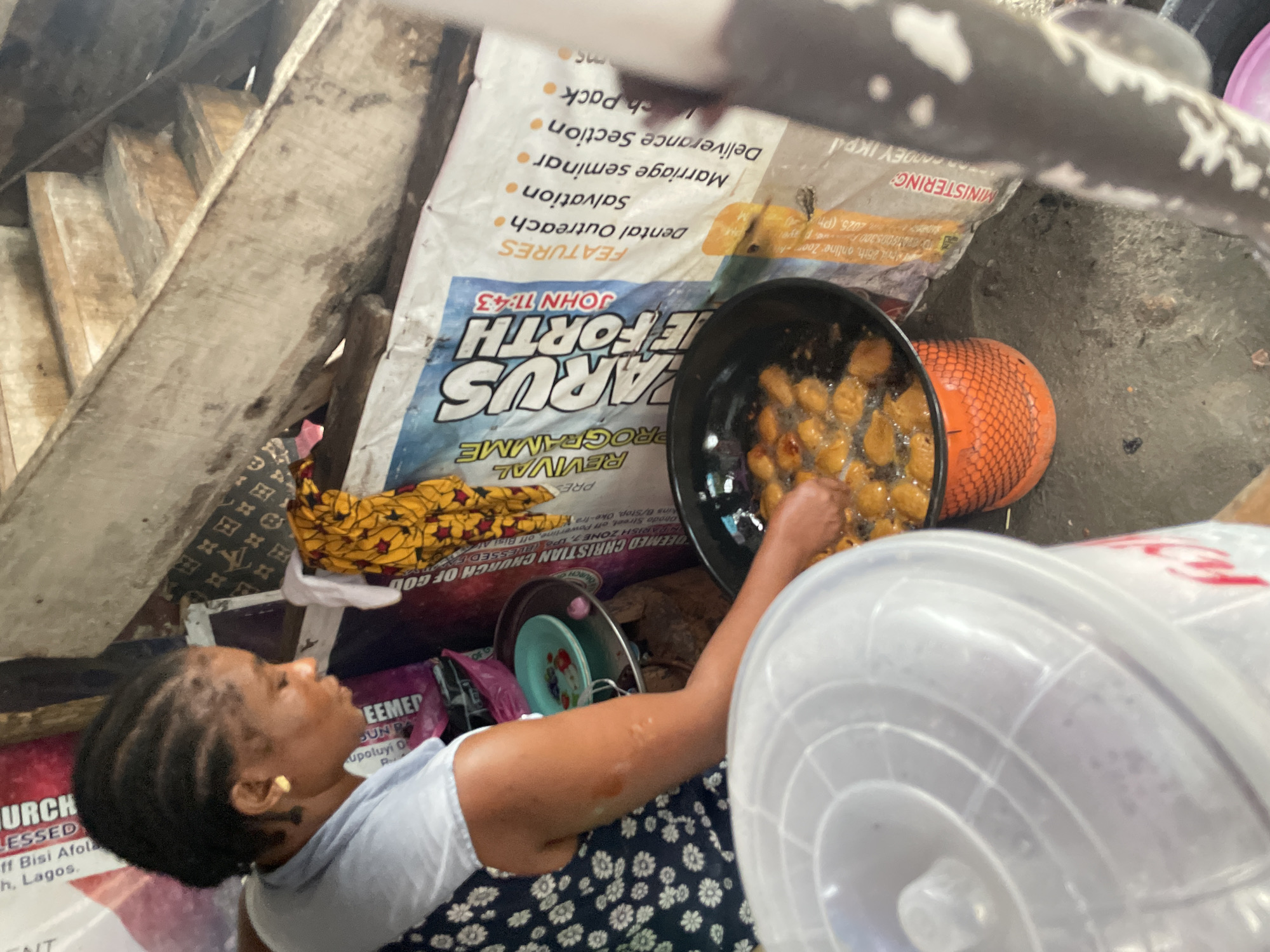 Here is a detailed description of the image:
**Scene:** An elevated, close-up view of a woman, likely in Ado, Nigeria, preparing and frying food in a market or similar informal setting.
**People:** A woman with braided hair is visible, wearing a light-colored top and a blue dress with a floral print. Her arm is extended as she tends to the food in a black cooking pot.
**Subjects/Objects:**
* A black cooking pot filled with oil and small fried dough items.
* An orange colander or strainer positioned near the cooking pot.
* Stacks of plates.
* Religious advertisement posters, including one for "PARUS EFORTH," featuring religious text like "JOHN 11:43" and "Salvation", with the church of focus being "REDEEMED CHRISTIAN CHURCH OF GOD." The address on the poster is partially visible as "Off Bisi Afolabi."
* A large, white plastic bucket or container dominates the foreground, partially obscuring the view.
**Actions/Interactions:**
* The woman is actively engaged in frying food.
**Environment:**
* The setting is cluttered and appears to be a temporary food preparation area.
* The backdrop consists of various items, including religious posters, and other cooking utensils, and what appears to be wooden stairs above.
* It has an urban setting, given the posters, and location is Ado, Nigeria.