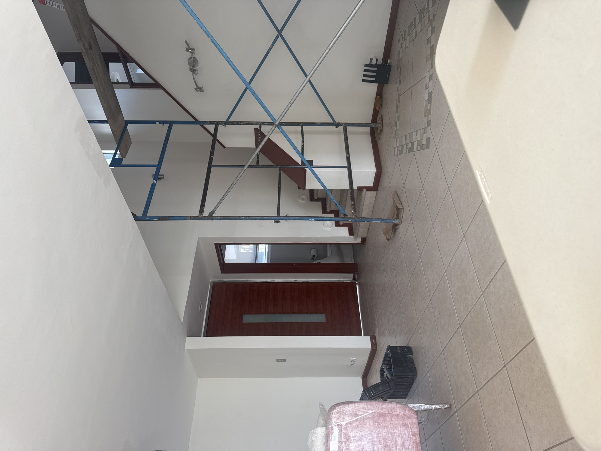 The image shows the interior of a building in Heroica Puebla de Zaragoza, Mexico, possibly a residential home. A temporary metal scaffold occupies the center of the frame, obscuring a set of stairs leading to an upper level. Below the stairs, there is a doorway with a dark wood door, partially open, revealing a glimpse of a bathroom. The floor is tiled with light brown, square tiles. Near the bottom of the frame, there are construction materials including what appears to be a wrapped bundle and an open toolbox. The walls are painted white.