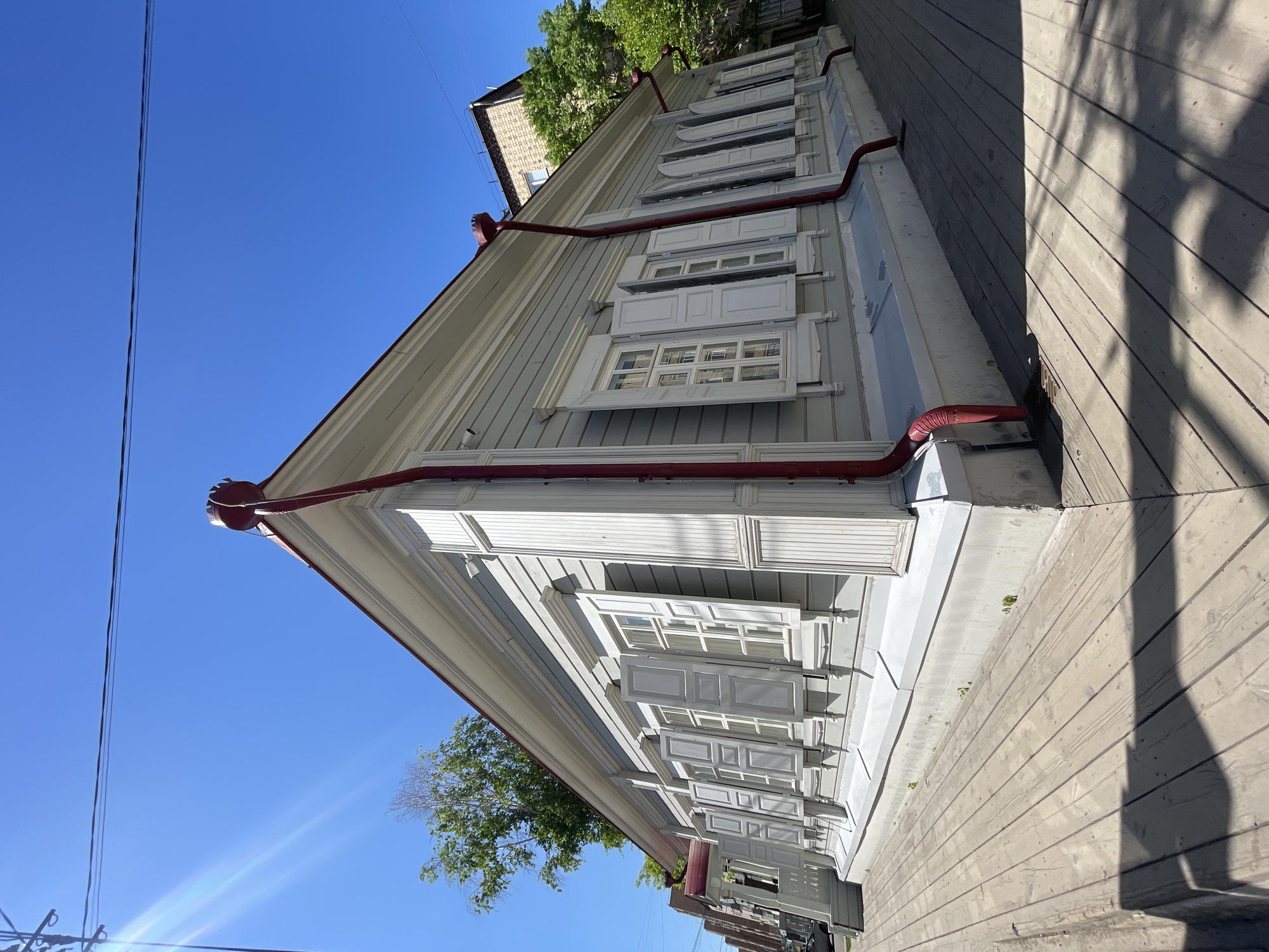 This image presents a picturesque view of a traditional wooden house, likely located in Krasnoyarsk, Russia, under a brilliant, clear blue sky. The house, extending diagonally from the foreground to the mid-ground, features light-colored (pale grey or white) wooden clapboard siding.
A prominent detail is the series of white-framed windows, each equipped with ornate, decorative white wooden shutters. Many of these shutters are partially open, showcasing their craftsmanship. Running along the eaves and corners of the house are vibrant red gutters and downspouts, providing a striking contrast against the pale walls and adding a pop of color to the structure. The house has a pitched roof, parts of which are visible, also adorned with the red guttering.
In the upper-left portion of the frame, thin utility wires stretch across the vast blue sky. Lush green trees, indicative of summer foliage, are visible behind the house, and beyond them, the tiled roof of another building can be discerned. To the far right, a wooden walkway or deck is bathed in bright sunlight, with long, distinct shadows cast upon its surface, suggesting the presence of nearby trees or other structures and indicating a clear, sunny day, likely mid-morning to afternoon. A subtle lens flare in the lower left corner further confirms the bright light conditions. The scene is devoid of people or activity, emphasizing the serene architectural beauty of the building. No visible text is present.