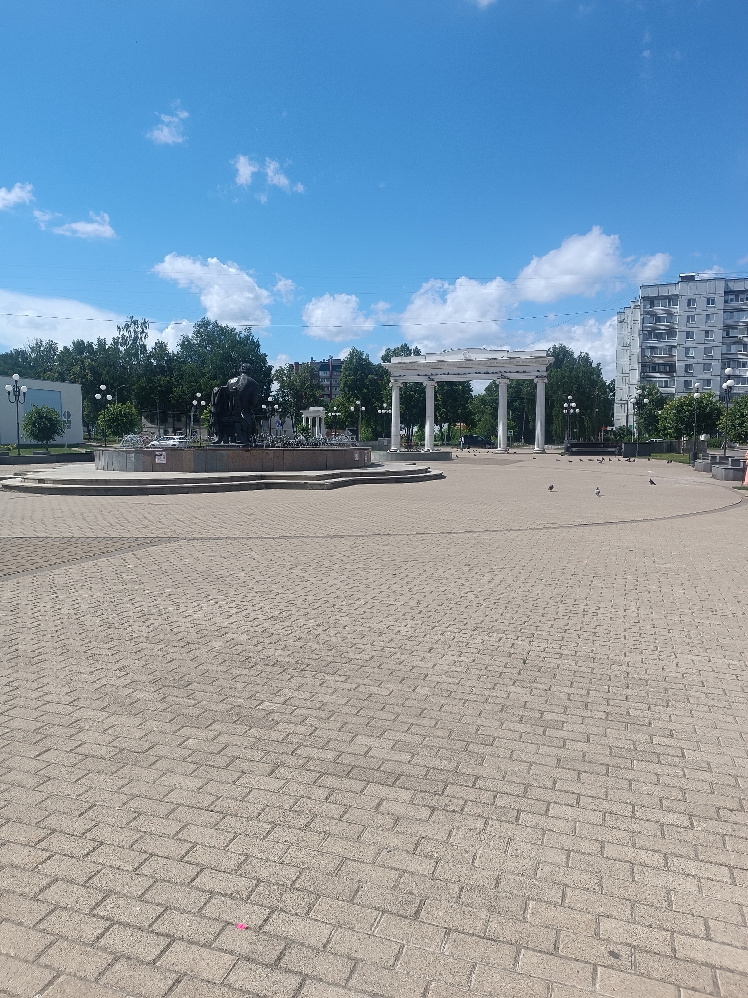 The image shows a wide, paved square with a sculpture and a fountain in the background. The square is made of light-colored brick pavers laid in a diagonal pattern. In the mid-ground, a large, dark sculpture of a seated figure is located within a tiered, circular fountain. The fountain appears to be inactive. Behind the fountain, a white, neoclassical-style archway with multiple columns stands. To the right of the archway is a tall, modern apartment building with a grey facade and numerous windows. Trees line the background, with a bright blue sky filled with scattered white clouds overhead. Several pigeons are scattered across the paved area. The scene appears to be a public space in the city of Chaikovsky, Russia, likely a central square or park.