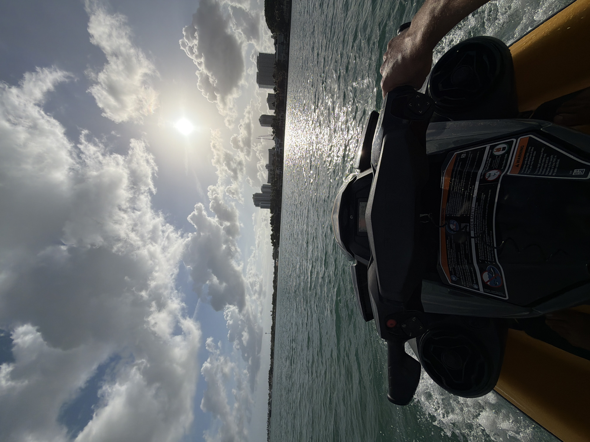 The image captures a water-level view from what appears to be a jet ski or similar personal watercraft, traversing calm water towards the skyline of Bal Harbour Village, United States of America. The sun is high in the sky, partially obscured by puffy white clouds, casting a bright glare on the water's surface. The jet ski's handlebars and part of the body are visible, along with a warning label providing rider safety instructions. The water is light green, and the city skyline is visible in the distance. A person's arm is also slightly visible.