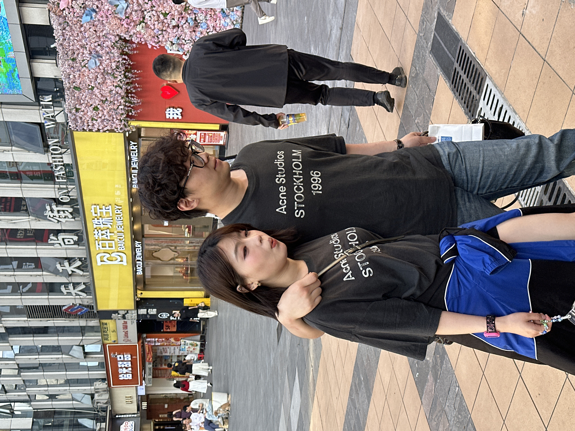 An outdoor urban street scene in Liangjiang, China, shows a young couple in the foreground, likely in a shopping district. The man, on the left, wears a black t-shirt, blue jeans, and glasses, with his arm around the woman's shoulder. The woman, on the right, also wears a black t-shirt, a blue and black jacket, and has dark hair, looking slightly upwards with a thoughtful expression. Both of their t-shirts read "Acne Studios STOCKHOLM 1996."
In the midground, a man in a black outfit walks away from the camera, holding a drink bottle. The background features buildings with various shop signs in Chinese and English, including "天天尚街 FASHION WAL", "白菜珠宝 BAICUI JEWELRY", "拾光甜品" (Shiguang Dessert), and "柯洛克密室" (Kolok Escape Room), alongside a prominent decorative wall covered in pink and blue artificial flowers. Other pedestrians are visible further down the street. The ground is paved with patterned tiles, and a large metal drain grate is present. Bright daylight illuminates the scene, suggesting it is daytime, possibly late afternoon.