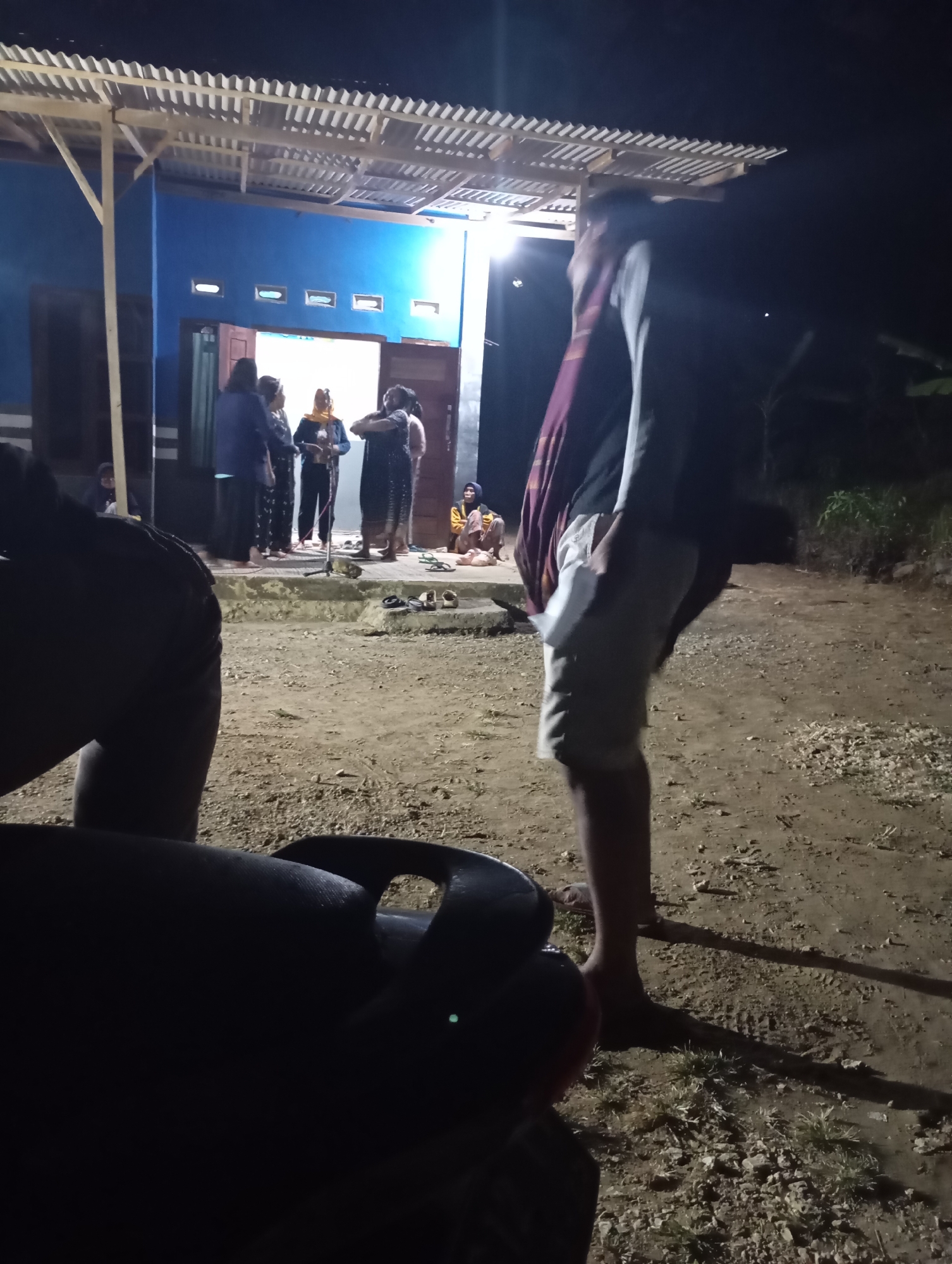 At night in Ketepung, Indonesia, several individuals are gathered outside a blue building. The building has a white, slatted awning extending from its roof and two windows on the upper section of the wall. A doorway is open, revealing a well-lit interior. Some people are standing in front of the doorway, appearing to converse. One person in the foreground is blurry and seen from behind, seated. The ground is dirt with scattered rocks and dry vegetation. A dark object, possibly a piece of furniture or equipment, is in the lower foreground, also out of focus.