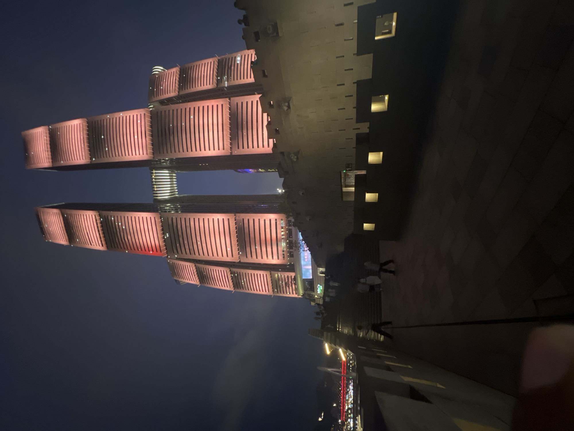 A nighttime, low-angle shot captures the iconic Raffles City Chongqing twin towers in Yuzhong District, China. The buildings are illuminated with a warm, pinkish-red light, highlighting their modern, segmented architecture. A distinctive horizontal skybridge connects the two towers, also lit with white lights. In the foreground, a textured, stone-like wall, reminiscent of ancient fortifications, runs along a pathway. Small, illuminated square openings are visible along this wall. Several people are walking on a wide, dark staircase leading up towards the towers. In the background, a bridge with red lights crosses a body of water, and distant city lights suggest a bustling urban environment. The sky is a deep, dark blue, with a few faint clouds.