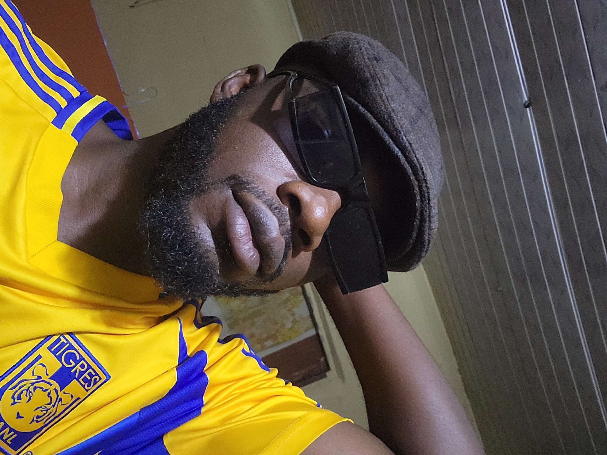 A close-up, waist-up portrait shows a Black man indoors, wearing a brown flat cap, black sunglasses, and a yellow and blue striped jersey. The jersey has a logo on the chest that appears to be that of the Tigres UANL football club. The man has a beard and is looking towards the camera with his right arm resting behind his head. The background is dimly lit and shows a textured ceiling with horizontal lines and a wall with yellow and brown tones. The image appears to have been taken in Jos, Nigeria.