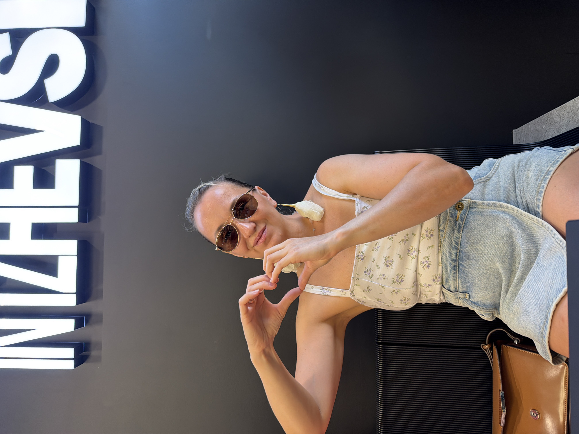 A woman with blonde hair, wearing sunglasses, a floral print top, and a denim skirt, is seated and making a heart shape with her hands in front of her chest. She is adorned with large, white, textured earrings. A brown leather handbag rests beside her on a dark, ridged surface. In the background, a large, illuminated sign displays the word "IZHEVSKI" in bold, white letters against a dark wall. The setting appears to be an indoor public space.