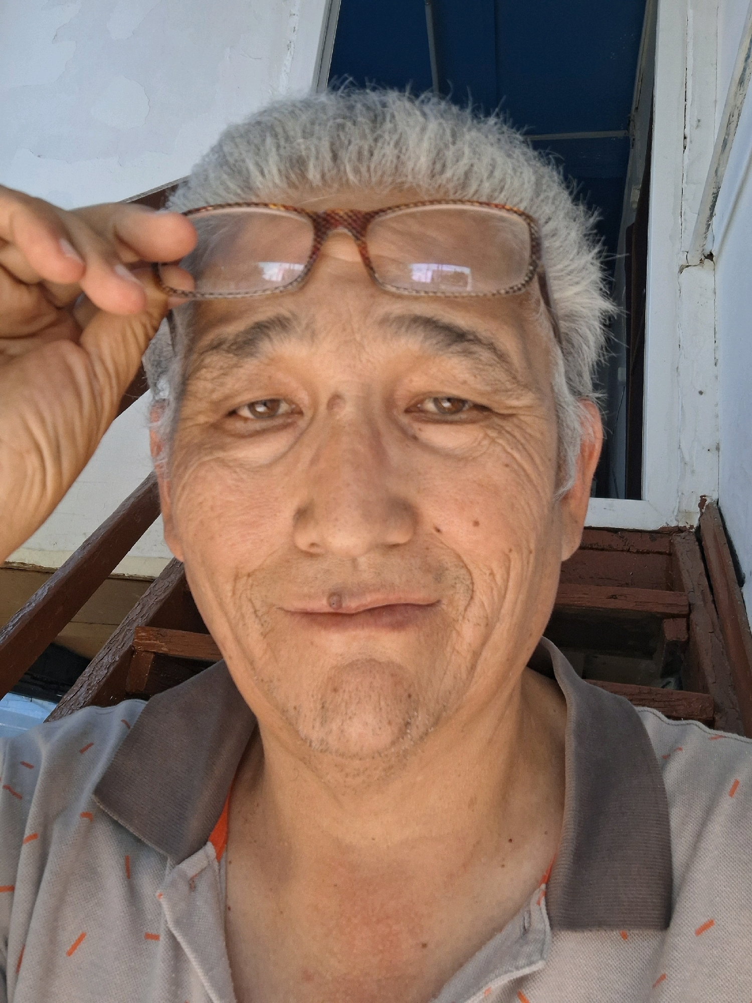 A close-up selfie taken outdoors shows a middle-aged man with gray hair and glasses perched on his forehead. He is wearing a light-colored polo shirt with a gray collar and orange accents. The background reveals weathered wooden stairs and a white wall, suggesting an older structure. The man's expression is neutral. The lighting indicates it is daytime. The scene is likely a residential area in Almaty, Kazakhstan, possibly a dacha or an older neighborhood with traditional architecture.