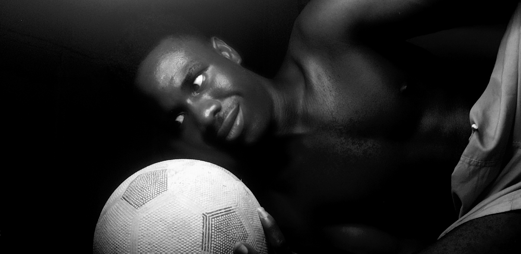 The image shows a shirtless dark-skinned man holding a soccer ball. He is lying down and looking at the camera with a slight smile. The background is dark, and the lighting is focused on the man and the ball. There is no other information in the image to associate it with Narayi, Nigeria.