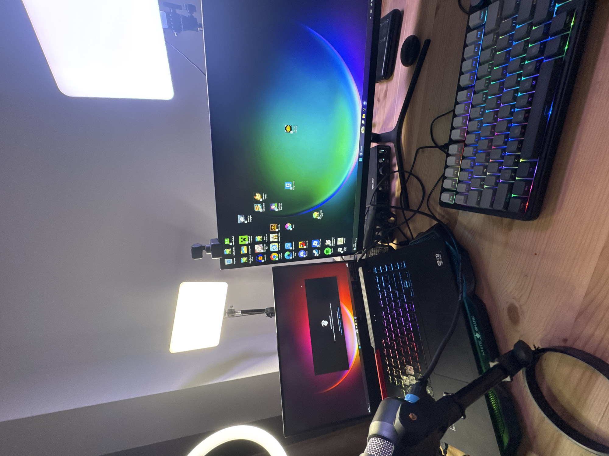 A cluttered desktop setup includes a large monitor displaying a screen with various icons, a laptop with an error message on its screen, and a mechanical keyboard with RGB lighting. A microphone and pop filter are also present. Additional lighting includes a ring light and two panel lights. The desk has a wood grain surface. The location in the city of Budva, Montenegro cannot be determined from the image.