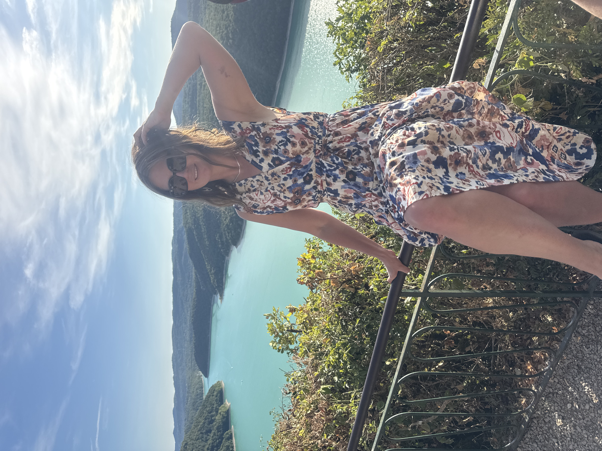 A woman is seated on a railing in front of a panoramic view of a turquoise lake surrounded by verdant hills. She is wearing a floral print dress and sunglasses, with her left hand resting on her head. The sun is shining brightly, casting shadows and highlighting the textures of the foliage and the water. The railing she is sitting on is made of dark metal with decorative curves. The background shows a vast expanse of water winding through the landscape, with a clear blue sky above. The image was taken in Moirans-en-Montagne, France, likely at a viewpoint overlooking the Lac de Vouglans.