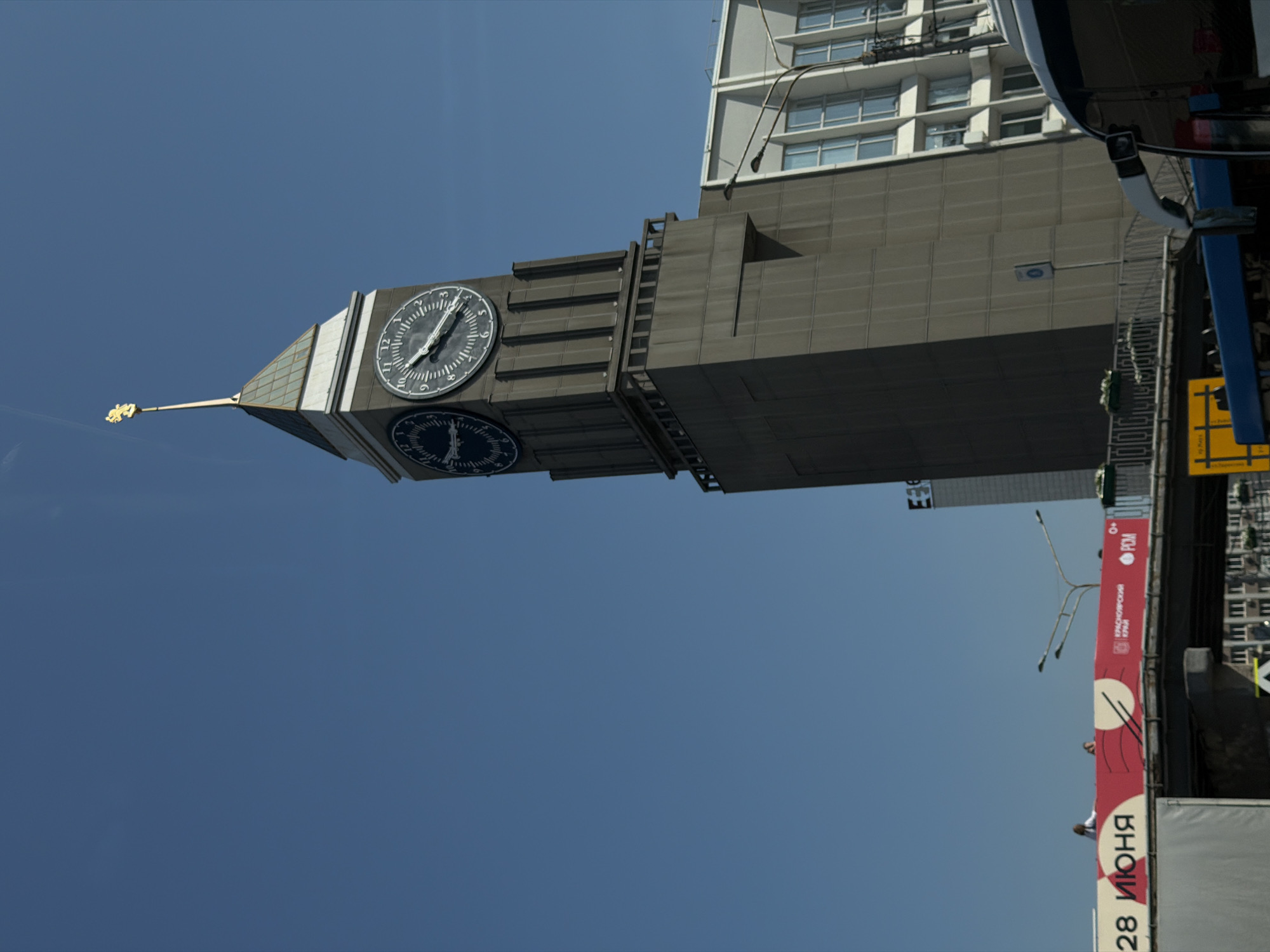 The image shows a view of the Krasnoyarsk Railway Station clock tower in Krasnoyarsk, Russia. The clock tower is tall and imposing, with two large clock faces visible. The sky is clear and blue. In the foreground, a red banner with text is visible, and the top of a bus is also in frame. The banner appears to advertise an event or date, with the text "28 ИЮНЯ" (June 28) prominently displayed. There are a few people visible on top of the banner structure. The architectural style of the buildings suggests a post-Soviet era construction.