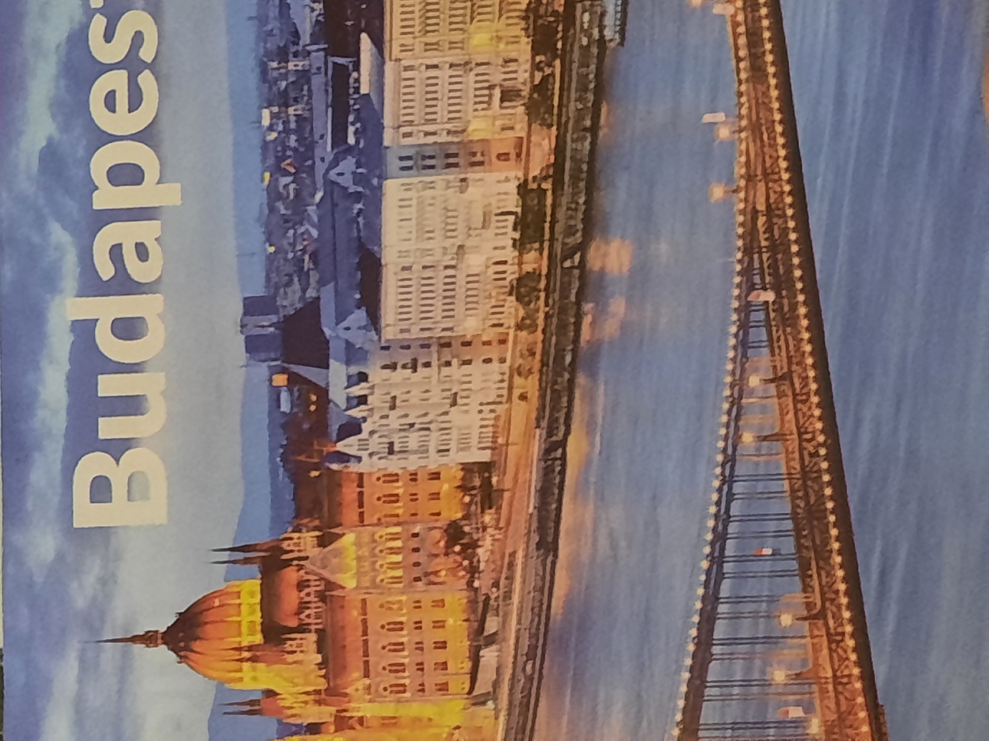 The image displays a postcard-like scene featuring a picturesque view of Budapest, Hungary, at dusk. The word "Budapest" is prominently displayed in large, white, sans-serif font across the top portion of the image. The scene itself is dominated by the illuminated Hungarian Parliament Building on the left, with its iconic dome and spires glowing warmly. Adjacent to it, a series of ornate buildings line the waterfront. In the foreground, the Chain Bridge stretches across the Danube River, its elegant structure adorned with numerous lights, creating a warm glow on the water. The river itself is a deep blue, reflecting the twilight sky. There is no indication of the city of Rosenheim, Germany in this image.