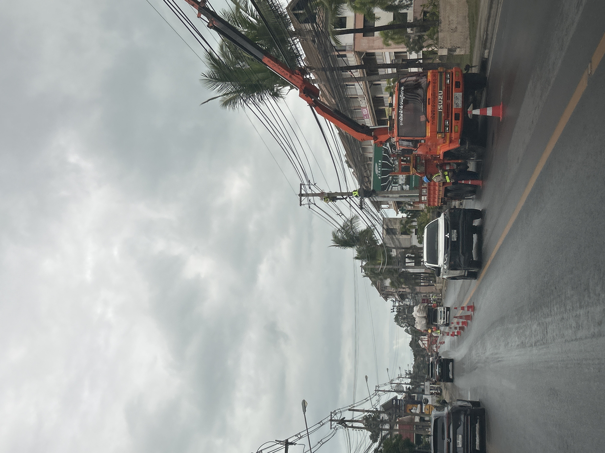 The image depicts a street scene in Ko Samui District, Thailand, under an overcast sky. The road is wet, suggesting recent rain. On the right side of the road, an orange Isuzu crane truck is parked, with its boom extended and resting on telephone poles. Two construction workers in yellow vests and blue helmets are near the truck. One worker is standing beside the truck, and another is visible on a pole, working on the electrical lines. Traffic cones are placed along the road, indicating ongoing work. Several vehicles, including SUVs and a truck carrying large bags, are present on the road, navigating around the work area. Buildings, some appearing to be commercial establishments and residential structures, line the street. Palm trees are also visible, characteristic of the tropical environment. The overall scene suggests a utility maintenance or repair operation in progress on a public street.