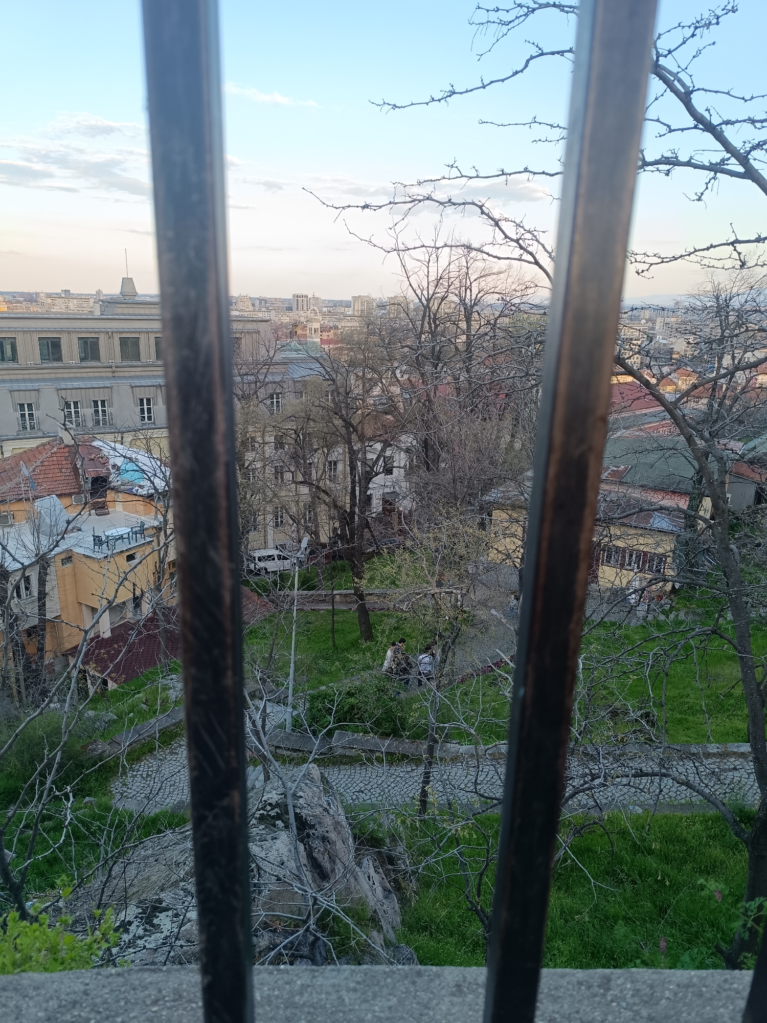 The image shows a view of a park and city skyline through wrought iron bars. The park features green grass, trees with bare branches, and a stone-paved pathway. Several people are seated on benches within the park. Beyond the park, a cityscape with buildings of varying heights and architectural styles stretches to the horizon. The sky is light blue with scattered clouds. The location is Plovdiv, Bulgaria, given the architectural characteristics of the buildings and the style of the wrought iron.