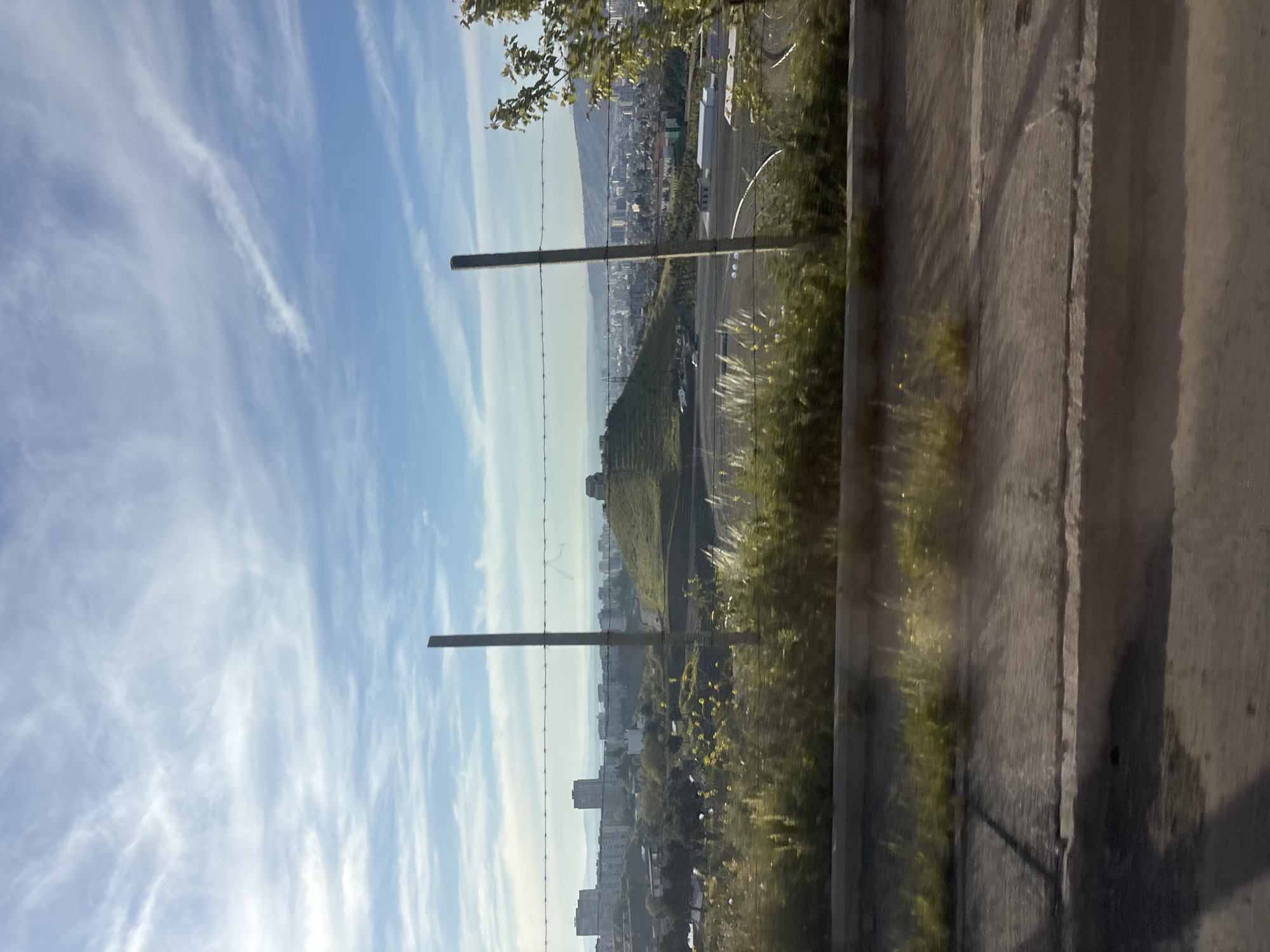 The image shows an elevated view of Tbilisi, Georgia, from near a concrete barrier topped with grasses. A barbed wire fence runs along the hill's edge. The sky is blue with wispy clouds. In the background is a city skyline. A green hillside with a structure near the top is also visible. The photograph appears to be taken at an angle, possibly from a moving vehicle.