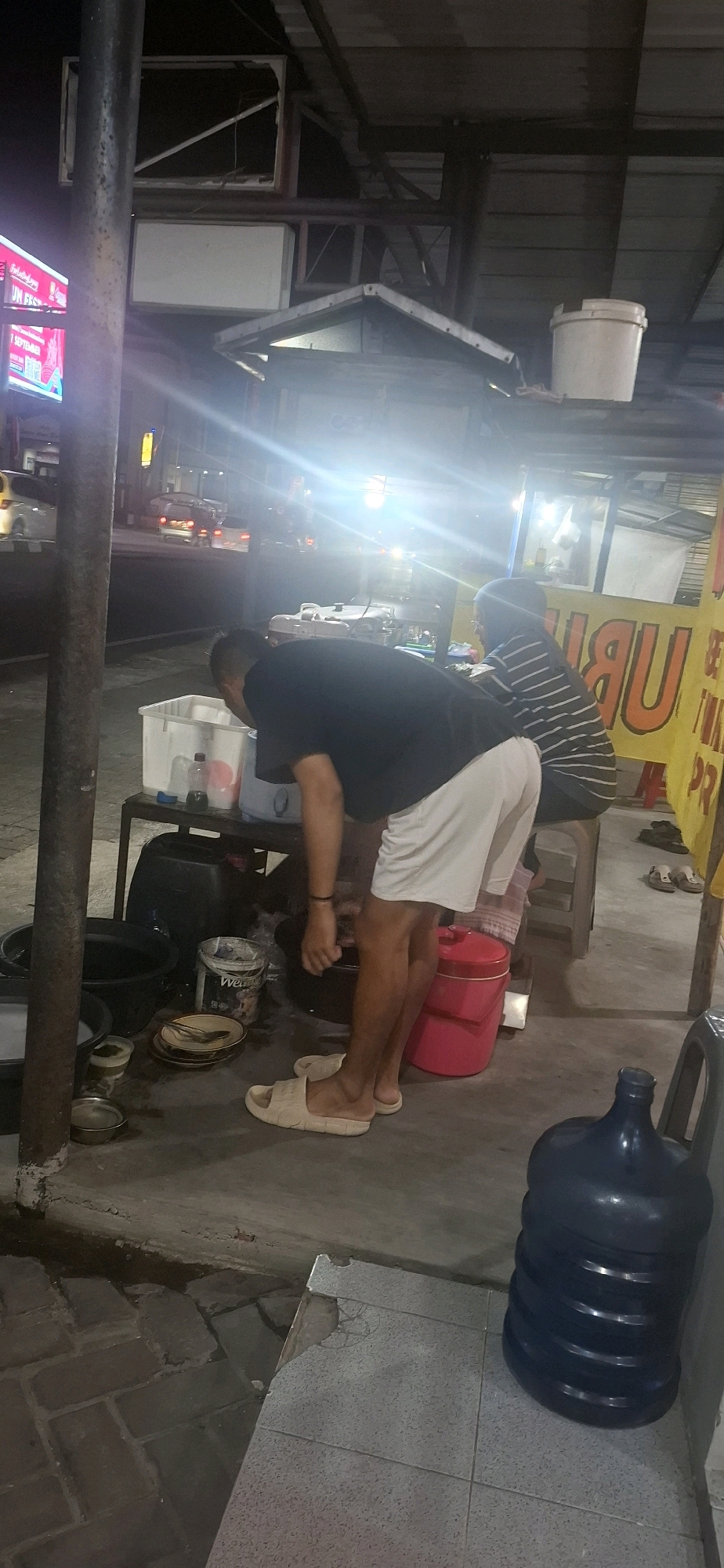 The image depicts an outdoor scene at night, possibly a street-side food stall, in Colomadu, Indonesia. A man wearing a black t-shirt and white shorts is bending over a table with various containers. He is facing away from the camera. Another person in a striped shirt is seated behind the table. In the foreground, there is a large dark blue water jug. The environment includes a concrete ground, a metal pole, and a structure with a tin roof. Bright lights from nearby sources create glare in the image. There are vehicles visible in the background on a street.