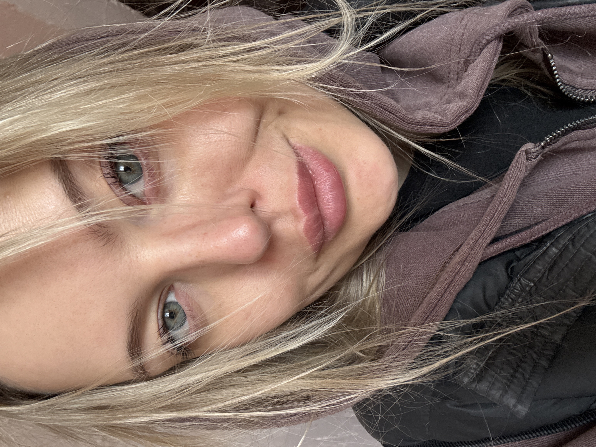 A close-up, diagonal portrait features a young woman with fair skin and light, possibly blue or grey-green, eyes looking directly at the camera. Her blonde hair, styled with highlights or layers, frames her face, with some strands falling across her forehead and eyes. She wears a slight, closed-mouth smile or smirk, conveying a relaxed and approachable emotion. Her lips are naturally full with a soft, pinkish hue.
The woman is dressed in what appears to be a purplish-brown hooded garment, possibly a hoodie or jacket, visible on the right side of the image. Beneath this, a black garment, potentially a jacket or an inner layer, is visible, featuring a zipper detail. The texture of the fabrics suggests casual wear.
The setting is an indoor, close-up shot, possibly a selfie, focusing on her face and upper body. The background is indistinct, appearing as a plain, light surface. The overall lighting is soft and even. There is no visible text in the image. This scene takes place in Borzya, Russia.