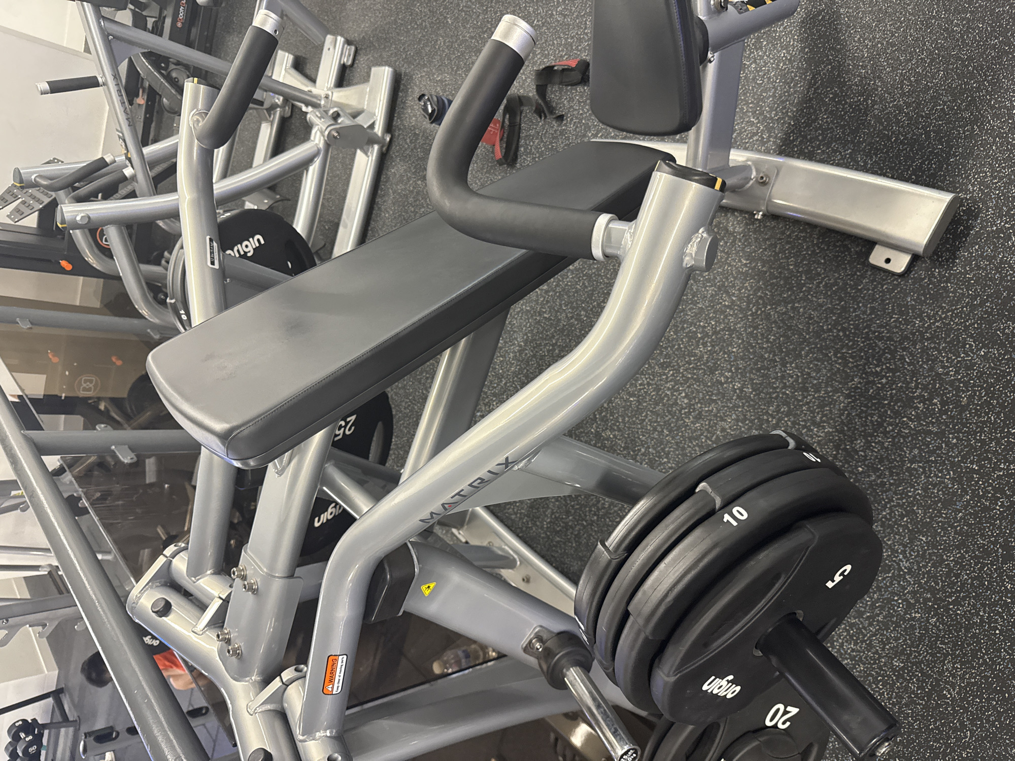 This close-up image captures a modern gym setting in London, United Kingdom, featuring a plate-loaded strength training machine. The scene is indoors, with a textured black speckled rubber floor typical of fitness centers.
The primary subject is a Matrix brand exercise machine, characterized by its sturdy silver-grey metallic frame and black padded seat and backrest. The machine's handles, designed for exercise, are covered with black rubber grips. Several black weight plates are loaded onto a weight horn attached to the machine. These plates are clearly marked with white numbers indicating their weight, including "20", "10", and "5", and many also bear the "origin" brand logo. An orange "A WARNING" label is visible on the machine's frame.
In the background, glimpses of other gym equipment, including another "25" weight plate and components of other machines, can be seen, hinting at a larger fitness facility. No individuals are present in the image, indicating no specific activity is taking place at the moment, but the equipment is set up for use. The lighting appears to be artificial, consistent with an indoor gym environment.