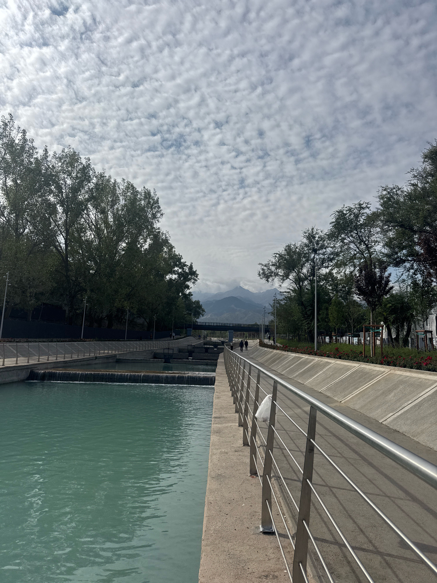 The image depicts a man-made canal in Almaty, Kazakhstan, likely part of the city's urban infrastructure designed to manage water flow and create recreational spaces. The canal itself is a prominent feature, with its turquoise-colored water flowing gently. Along the canal's edge, a paved walkway is visible, separated from the water by a concrete embankment. A sleek, modern metal railing lines the walkway, suggesting a well-maintained public area.
On the walkway, two individuals are seen walking away from the camera, their figures silhouetted against the brighter background. They appear to be enjoying a stroll along the canal. To the left of the canal, lush green trees with full foliage line the bank, creating a natural buffer. In the distance, a bridge can be seen spanning the canal, and beyond that, a range of mountains rises against a cloudy sky. The sky is filled with a pattern of white, cumulus clouds, interspersed with patches of blue.
The overall impression is one of a serene urban landscape that blends natural elements with modern infrastructure. The presence of trees, water, and mountains suggests a city that values its connection to the natural environment. The walkway and railing indicate that this area is designed for public use and enjoyment, likely a popular spot for residents and visitors alike to engage in outdoor activities. The scene is peaceful, with no apparent signs of congestion or activity beyond the two walkers.