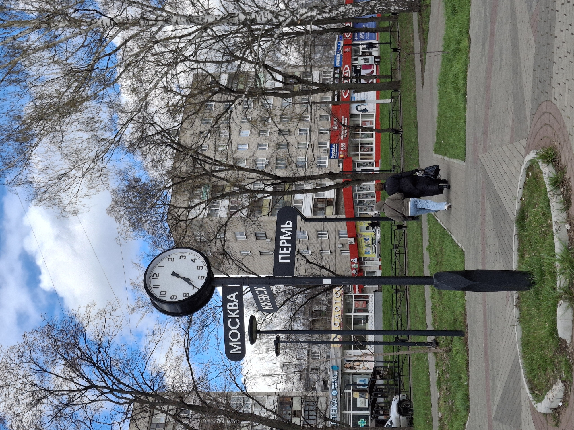 This image captures an urban street scene in Chaikovsky, Russia, likely during late autumn or early spring, given the bare branches of the trees. The time is approximately 4:35, as indicated by a prominent black street clock in the foreground.
Two people are seen walking away from the camera on a paved sidewalk. One individual wears a dark coat and carries a bag, while the other wears a lighter jacket and blue jeans. To the left of the path, a black signpost features directional signs pointing to various Russian cities, clearly labeled "МОСКВА" (Moscow), "ИЖЕВСК" (Izhevsk), and "ПЕРМЬ" (Perm) in white Cyrillic text.
The foreground consists of a grey paved sidewalk with a reddish-brown brick pattern, alongside strips of green grass. A low black metal fence runs parallel to the sidewalk. In the background, behind the bare trees, multi-story buildings line the street. Many of these buildings appear to house ground-floor businesses with colorful signs. Visible text on these signs includes "КЛАСС" (Klass), "Pedant.ru", and partial words like "ТЕКА ЗДОРОВЬЯ" (likely part of "Pharmacy of Health") and the number "585". The sky above is partly cloudy with significant patches of blue. A white car is partially visible near the bottom left, parked alongside one of the buildings.