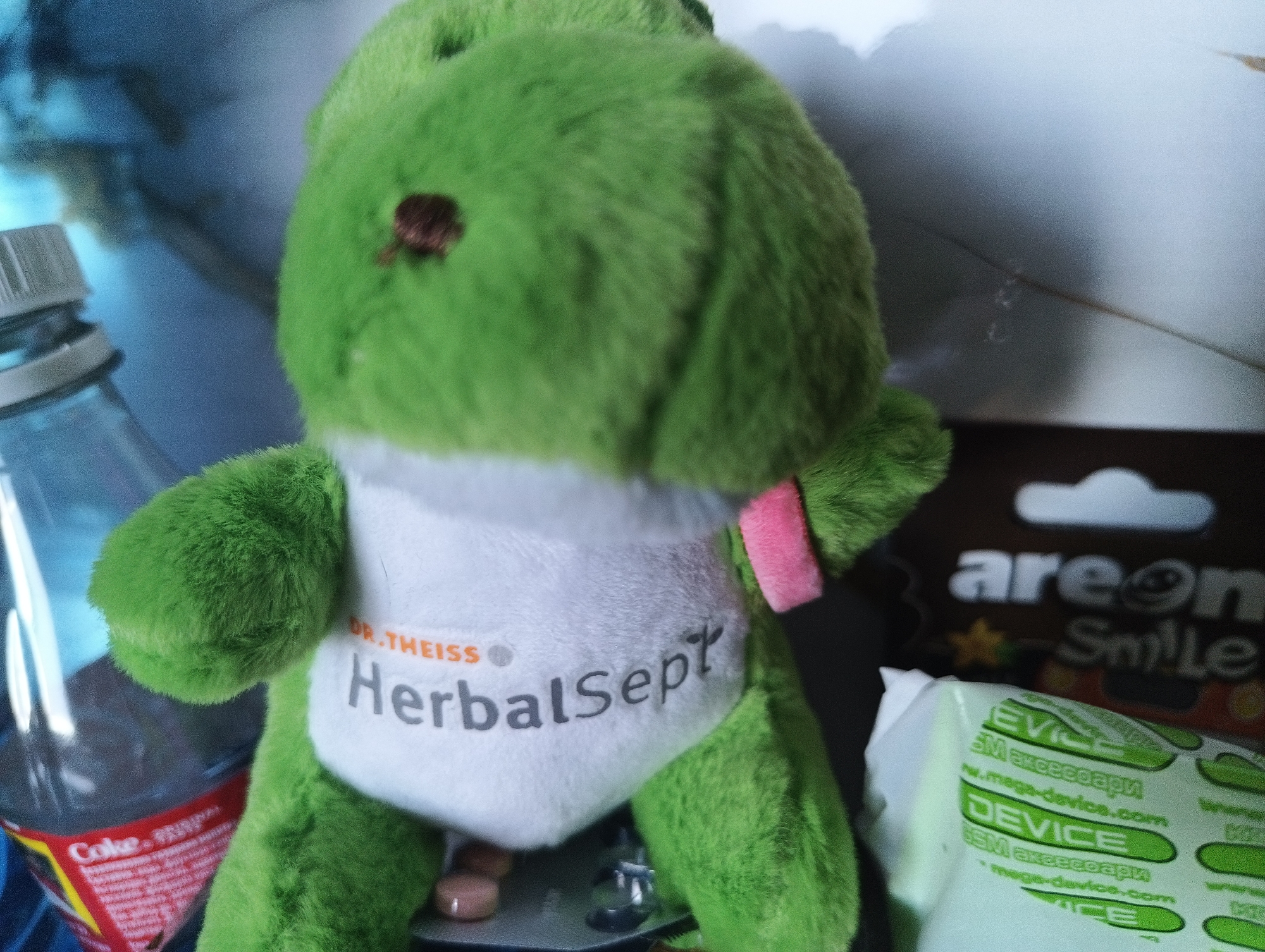 The image shows a close-up of a green plush toy, possibly a frog or dinosaur, with a white bib or apron. The bib has text that reads "DR.THEISS HerbalSept" in orange and gray letters. To the left of the toy is a clear plastic bottle with a red label that appears to be a Coca-Cola bottle. The bottle is partially obscured by the plush toy. In the background, to the right of the toy, there is a dark rectangular object with white text that reads "areon Smile". Below this, there is a white packet with green text that says "DEVICE". The overall scene is indoors, on a surface that is likely a table or shelf. The lighting is diffused, suggesting an indoor environment. The plush toy is the central focus of the image, positioned in the foreground. There are no people visible in the image. The image is a still life composition, and there is no discernible action or interaction taking place.