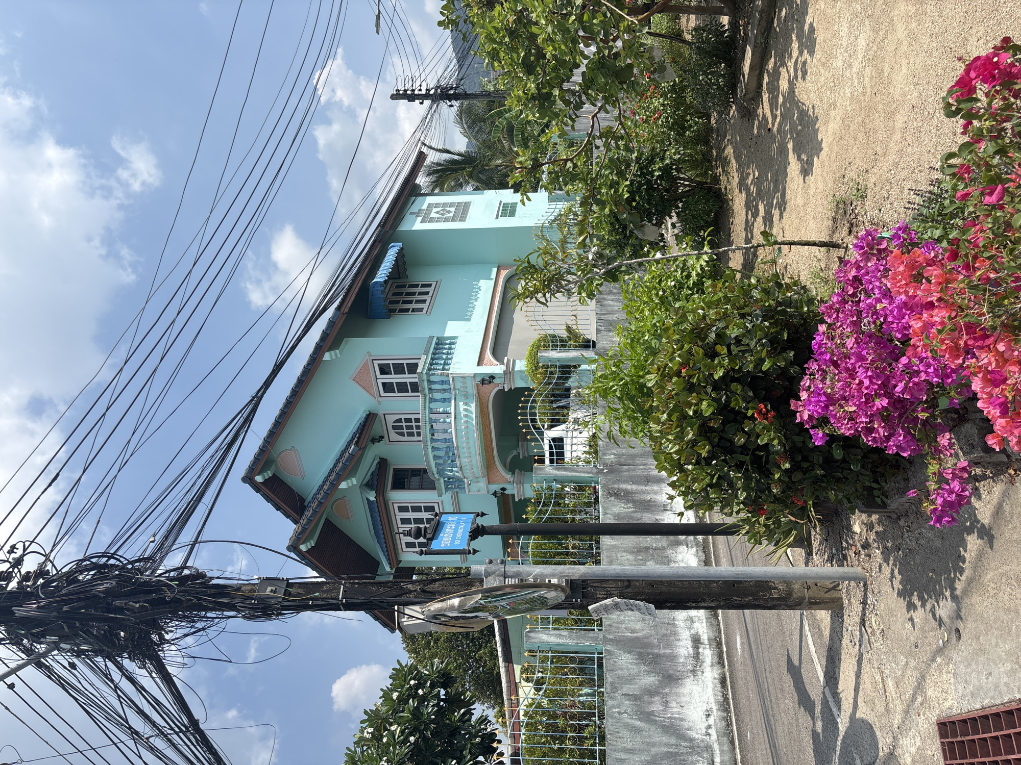 An outdoor, daytime photograph showcases a vibrant street scene in Choeng Thale, Thailand. The primary subject is a two-story house painted in a distinctive turquoise or light blue hue, featuring white window frames and intricate white railings on its balconies and fences. A blue awning extends over one of the ground-floor windows.
In the foreground, a prominent utility pole dominates the left side of the frame, laden with a dense, tangled mass of black electrical and communication wires. Further along the street, another pole supports a blue street sign and a convex traffic mirror. The street sign displays Thai script "ซอยช่างหล่อ 10" (Soi Changlor 10) and English transliteration "SO1 (A) CHERNGTALAY," confirming the location context.
The scene is bathed in bright daylight, evidenced by clear shadows cast on the ground, suggesting a sunny day with a few scattered white clouds visible in the blue sky. Lush green foliage and bushes line the roadside and surround the house. On the right, a large bush bursts with clusters of bright pink and purple Bougainvillea flowers, adding a splash of color to the scene. The ground transitions from a dirt or gravel path to a paved road, partially visible on the right. A concrete wall and a metal grate are also present on the right side of the image. No people or specific activities are depicted; the image captures a static, residential street view.