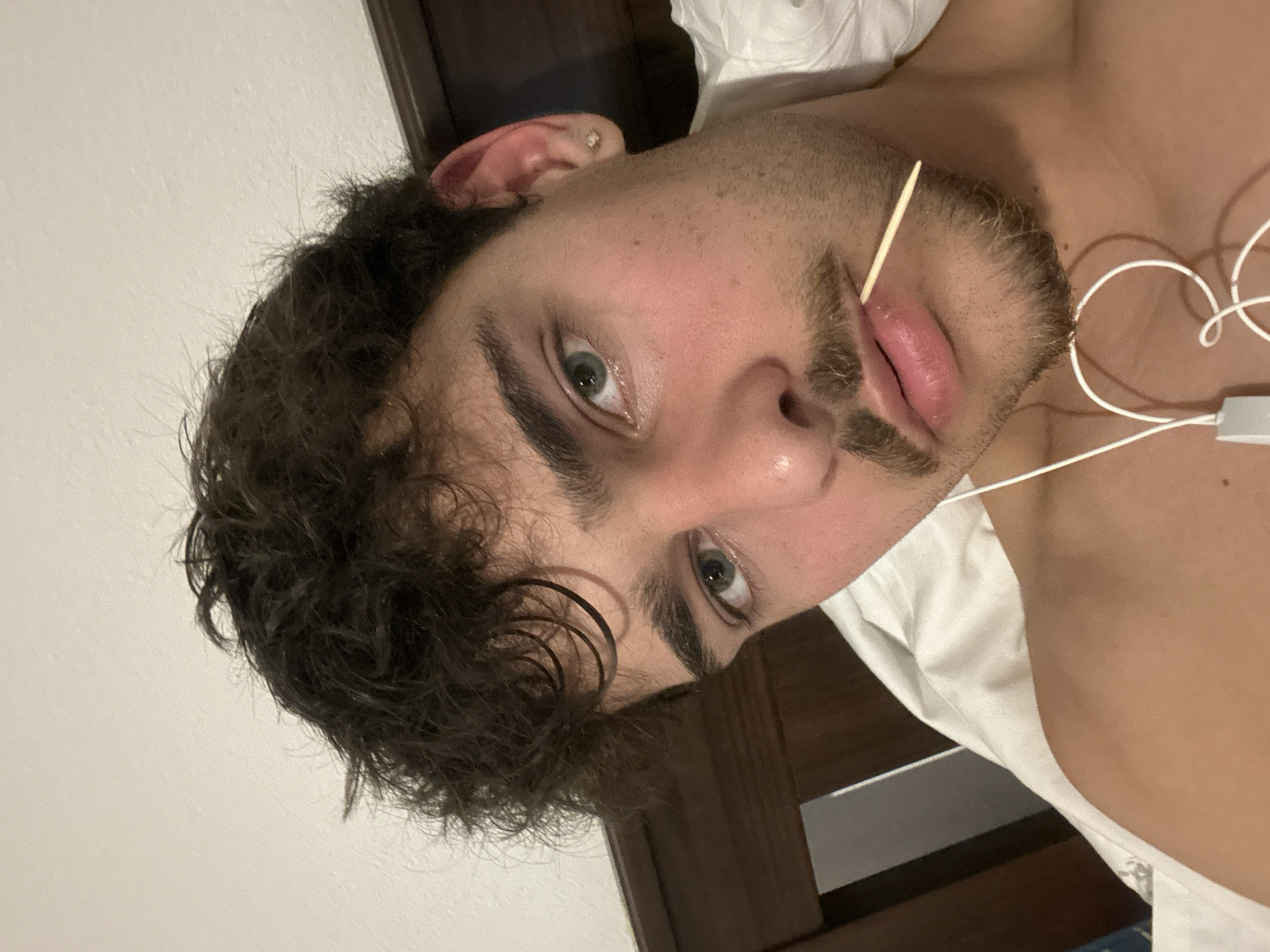 A selfie angled upwards shows a light-skinned young man lying on what appears to be a white bed sheet. He has dark curly hair, green eyes, and a mustache and beard. A thin wooden stick protrudes from the corner of his mouth. White wired earbuds are draped across his chest. A dark wooden door frame is visible in the background. The location of the image in the city of Troyan, Bulgaria, cannot be determined from the image.