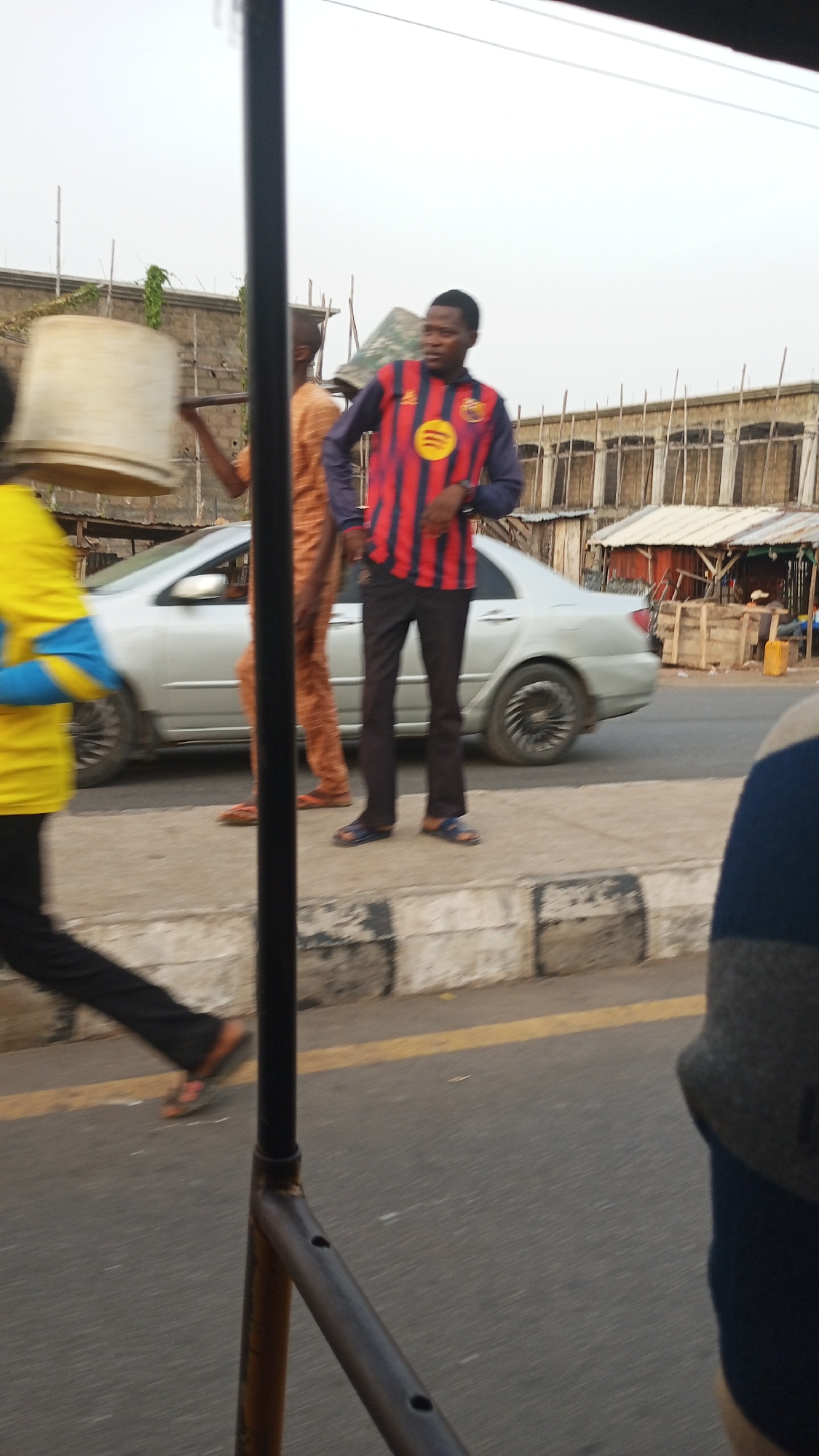 The image shows a street scene in Zango-ndaji, Nigeria. A silver car is parked on the side of the road. Three people are standing on the sidewalk. The person closest to the camera is wearing a red and black striped shirt with a circular yellow logo. Another person is wearing a patterned orange jumpsuit. A third person is carrying a white bucket above their head. Other buildings and structures are visible in the background. The scene appears to have been shot from inside a moving vehicle.