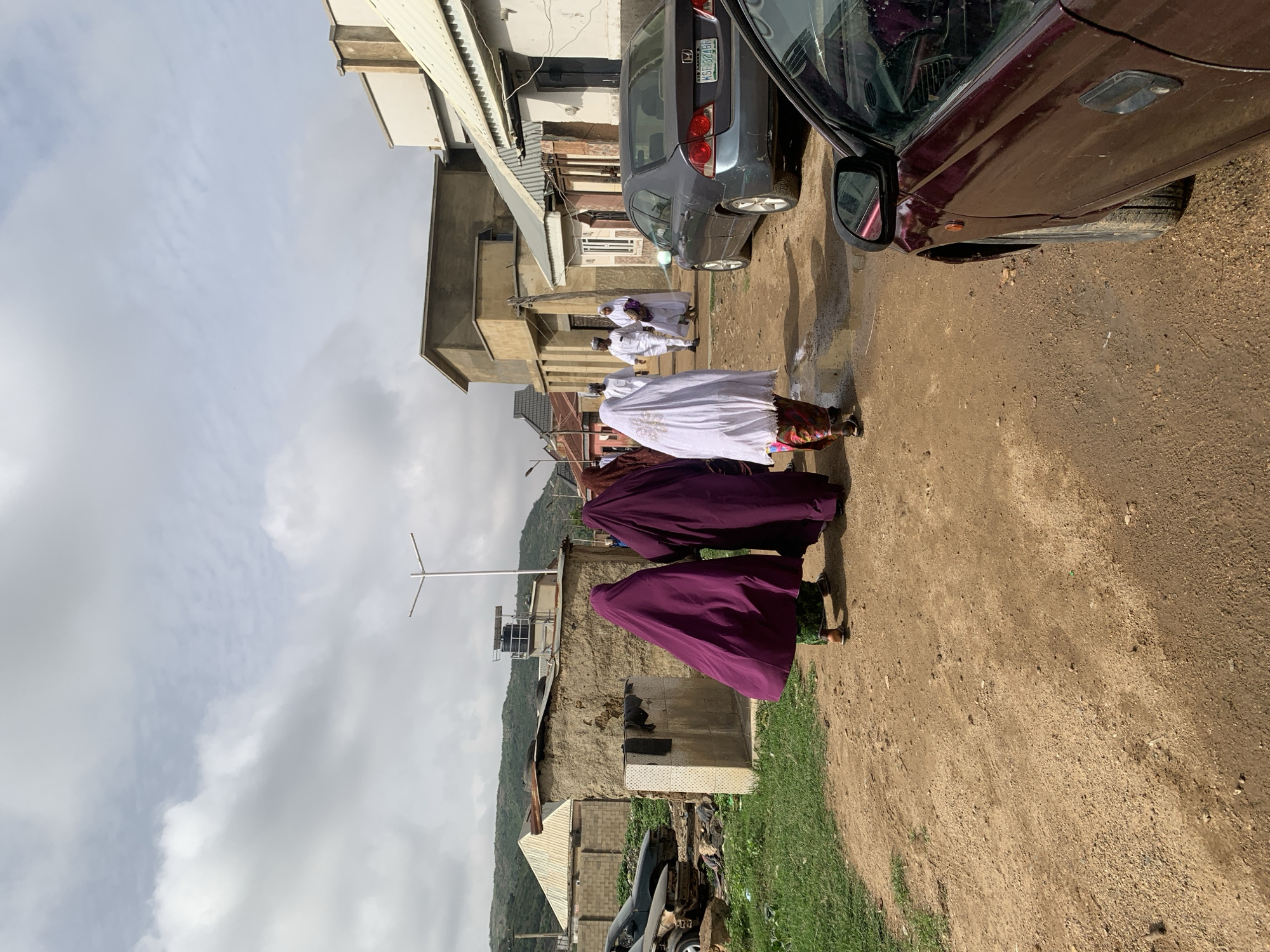 The scene is an unpaved street in Naraguta Hausawa, Nigeria. Two cars are parked on the right side of the frame. Four people are walking on the street; two are wearing full-length purple garments, and two are wearing white garments. A building is on the left side of the frame, with a mountain range in the background. The sky is overcast.