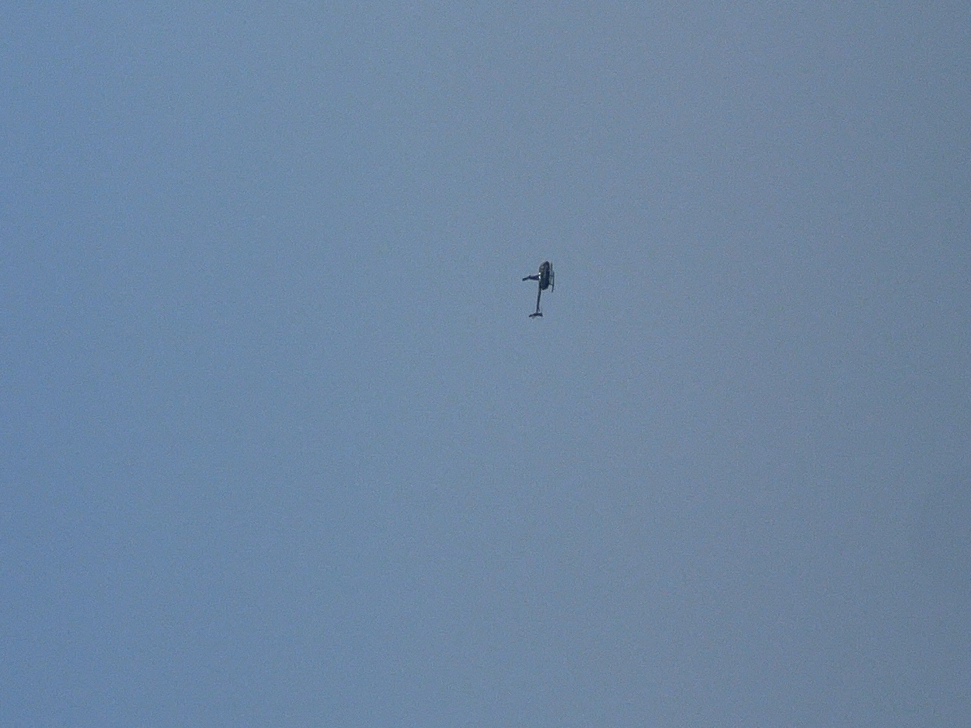 A single, dark-colored helicopter is captured in mid-flight against a clear, pale blue sky. The helicopter is positioned in the lower right quadrant of the frame. It is oriented with its nose pointing slightly to the right and appears to be a small, two-bladed model. The environment is entirely sky, with no discernible ground features, buildings, or other elements of Kuta, Indonesia visible. The image lacks any signs of human activity beyond the helicopter's presence.