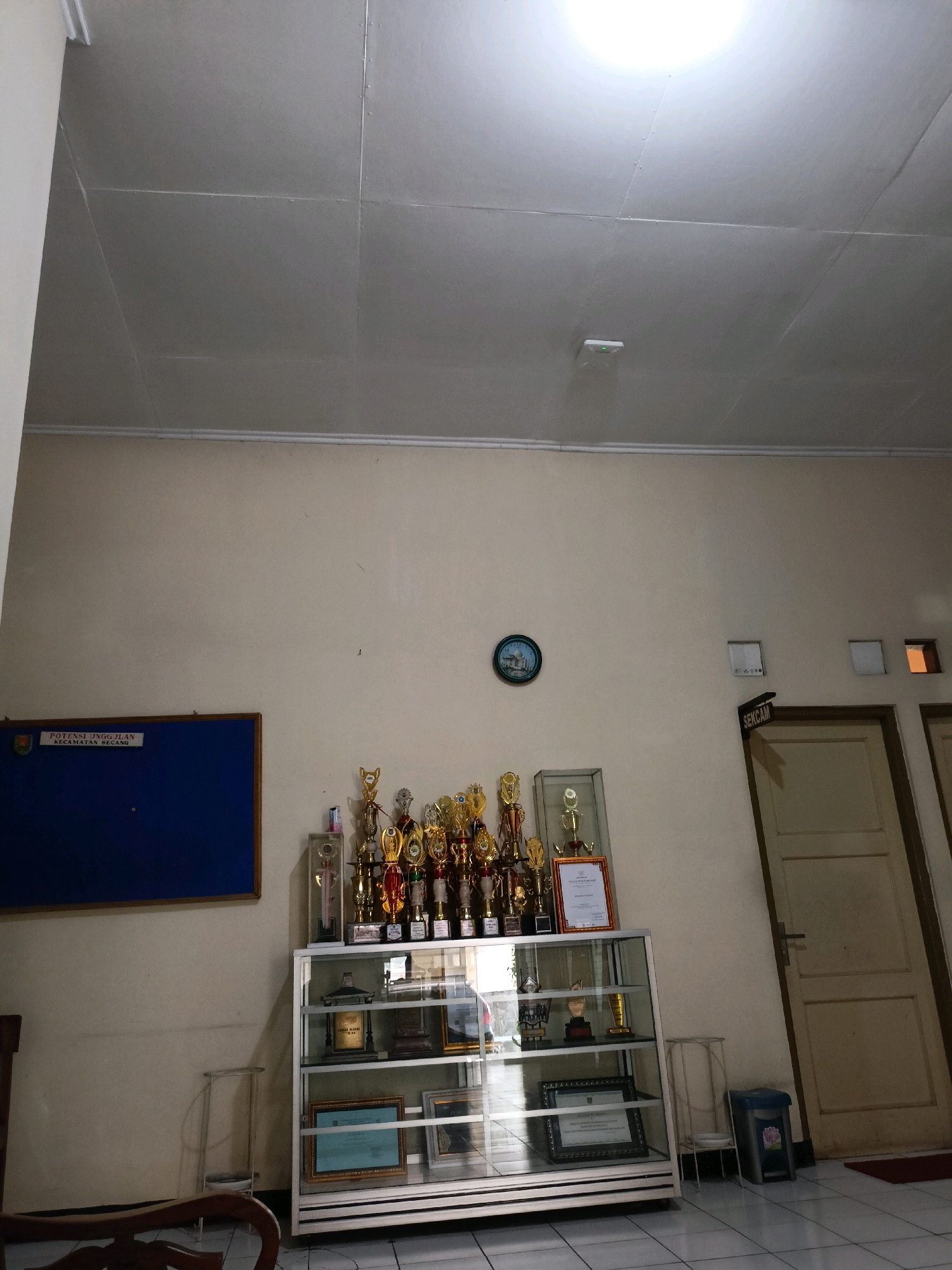 The image depicts an indoor scene, likely an office or reception area, with elements suggesting an institutional setting in Kewaluhan, Indonesia. A pale yellow wall dominates the background, with a white, paneled ceiling above. To the left, a dark blue bulletin board with a wooden frame is mounted on the wall, displaying text that reads "POTENSI UNGGULAN KECAMATAN SEKARAN". Below the bulletin board and slightly to the right is a wooden chair.
In the center of the image, a glass display cabinet stands filled with trophies, awards, and framed certificates. The trophies, mostly gold and silver, are arranged on the top shelf, while the lower shelves contain framed items, one of which appears to be a certificate with blue and white text, and another framed document with a crest. To the right of the display cabinet, a wall-mounted clock with a circular face and Arabic numerals is visible. Above the clock, there are two square white panels on the wall, and to the far right, a wooden door with a dark handle is present. A sign that reads "SEKCAM" is affixed to the wall above the door.
Further to the right, another wooden door is visible, slightly ajar. A blue trash can with a pink flower motif is placed on the floor near the doors. The floor is tiled with white squares. The lighting in the room appears to be artificial, with a bright light source visible on the ceiling. The overall impression is one of a functional space that also serves to showcase achievements and information.