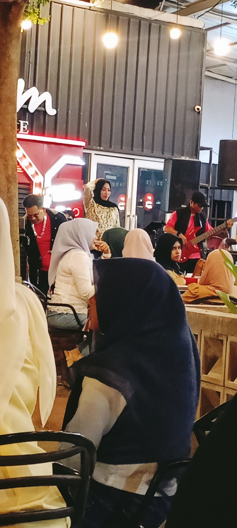 Captured in Lhokseumawe, Indonesia, the scene unfolds at a cafe. Patrons, predominantly women wearing hijabs of varying colors, are seated. A live band provides entertainment; a male guitarist, dressed in a red shirt and black vest, plays. A standing woman in a patterned blouse adjusts her hijab. Architectural elements include a dark gray, possibly metal facade, lights, and furniture typical of a cafe setting.