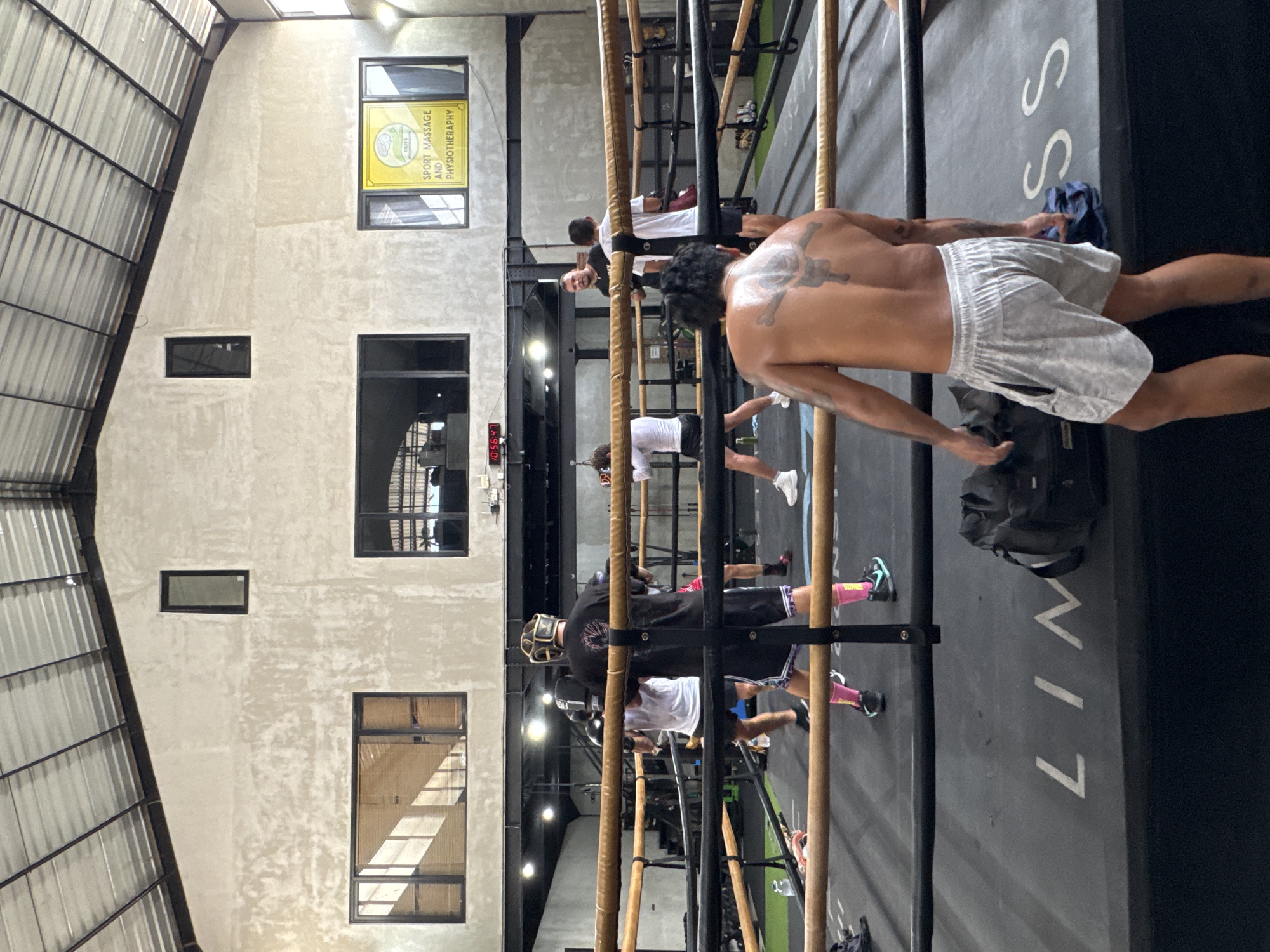 The image captures an active indoor fitness facility in Mengwi, Indonesia, characterized by its industrial aesthetic with light grey, textured concrete walls and a high ceiling featuring corrugated metal panels. Several black-framed windows punctuate the wall, allowing natural light into the space.
In the foreground, a shirtless man with prominent tattoos across his shoulders and upper back stands, wearing light grey athletic shorts. He appears to be resting or observing the activity, with a dark gym bag placed on the dark grey floor beside him. The floor features white markings, including partial text that reads "LIV" and "SS".
Further into the background, other individuals are engaged in training, positioned among a structure of vertical wooden poles and horizontal metal bars, suggesting a functional training or calisthenics area. One participant in a black long-sleeved shirt is visible wearing protective headgear while interacting with a pole. Another person in a white t-shirt is also part of the group. A digital clock mounted on the far wall clearly displays the time as "10:56:47".
A bright yellow sign is prominently displayed in an upper window, bearing the text "SPORT MASSAGE AND PHYSIOTHERAPY," alongside a logo that appears to include the word "CANGGU" or "CANG." This indicates the presence of recovery services, potentially located within or nearby the facility. The scene depicts a mid-morning workout session, reflecting a dynamic and health-focused environment typical of a gym or training center.
