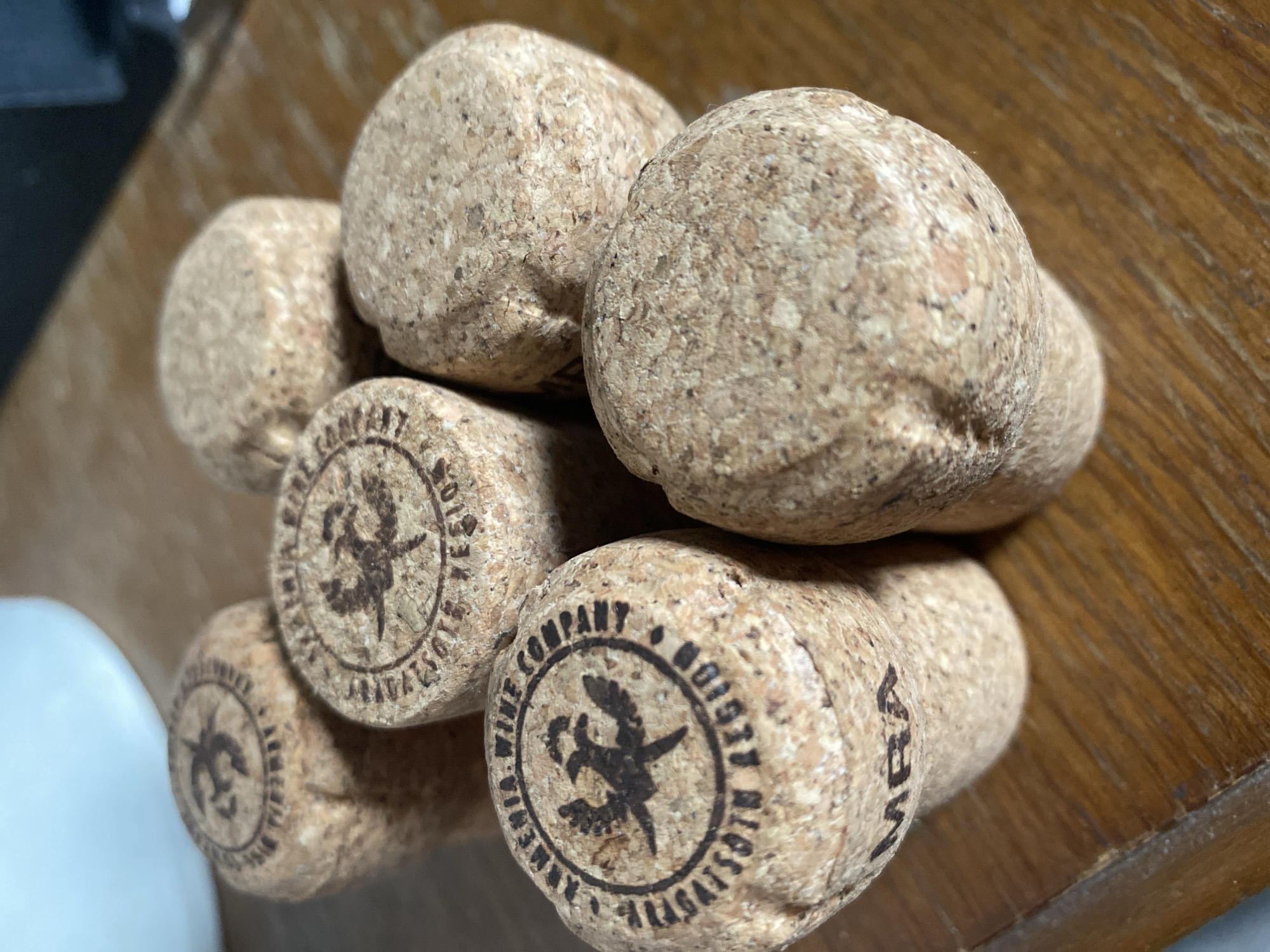 Six champagne corks are arranged in a cluster on a wooden surface. Each cork is cylindrical with a rounded top and has text and an emblem stamped onto it. The text on some of the corks reads "ARMENIA WINE COMPANY" and "ARAGATSOTN REGION". One cork also has the letters "MRA" stamped on its side. The wooden surface appears to be a table with visible grain. The background is blurred, but suggests an indoor setting with a white object and a dark, possibly electronic device. The lighting is natural, coming from the left, casting shadows on the corks and the table. The image is taken from a slightly high angle, looking down on the corks. The location in Им. Коняшина Снт, Russia is not determinable from the visual information provided.