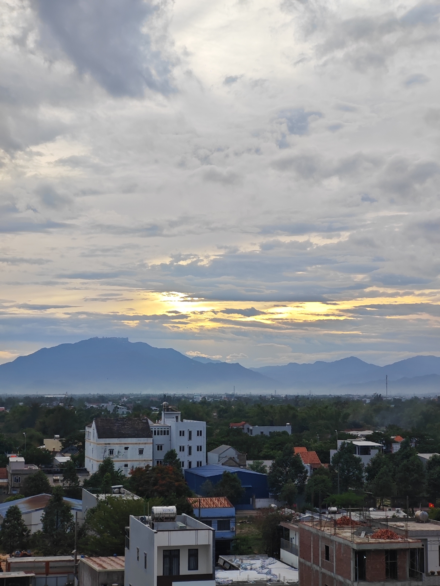 This image captures a wide-angle view of a city, likely Đà Nẵng, Vietnam, during what appears to be either sunrise or sunset. The sky is dominated by a dramatic arrangement of clouds, with patches of bright sunlight breaking through in the middle, illuminating the horizon.
In the distance, a prominent mountain range stretches across the landscape, its peaks softened by a light haze. Below the mountains, a sprawling city can be seen, characterized by a dense collection of buildings of varying sizes and architectural styles. Many buildings are residential, with pitched roofs and multiple stories. Some construction sites are evident, with exposed brickwork and scaffolding. Dense green foliage, likely trees and vegetation, intersperses the urban structures, suggesting a city with ample greenery.
Several distinctive buildings stand out within the cityscape. One notable structure is a white, multi-story building with a somewhat dated appearance, featuring many windows. Another building in the foreground appears to be under construction, with unfinished brick walls and visible rebar.
The overall atmosphere is one of tranquility and a sense of an expansive, developing urban environment juxtaposed with natural geological features and atmospheric conditions. There are no visible people or specific actions being depicted. The scene is static, offering a broad overview of the city and its surroundings.
