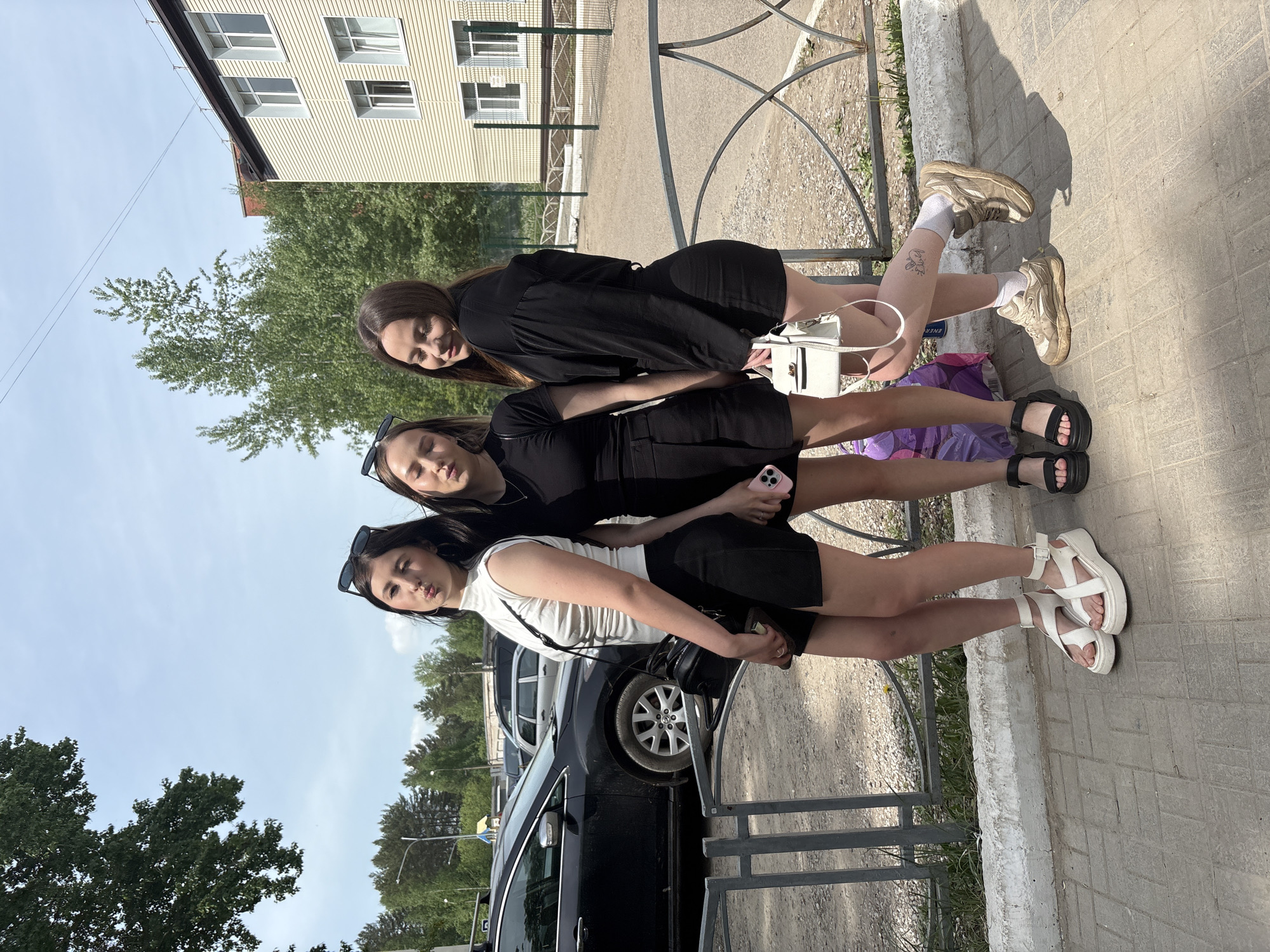 Three young women are standing in front of a low metal fence on a sidewalk, with parked cars and trees visible in the background. The woman on the right is wearing a black dress and tan sneakers with white socks. She has a small tattoo on her right leg. The woman in the middle wears a black dress and black sandals, holding a pink phone. The woman on the left is wearing a white top, black skirt, and white sandals. They appear to be posing for a photo. The image was taken in the city of Chaikovsky, Russia. A multi-story building with light-colored siding is visible in the background.