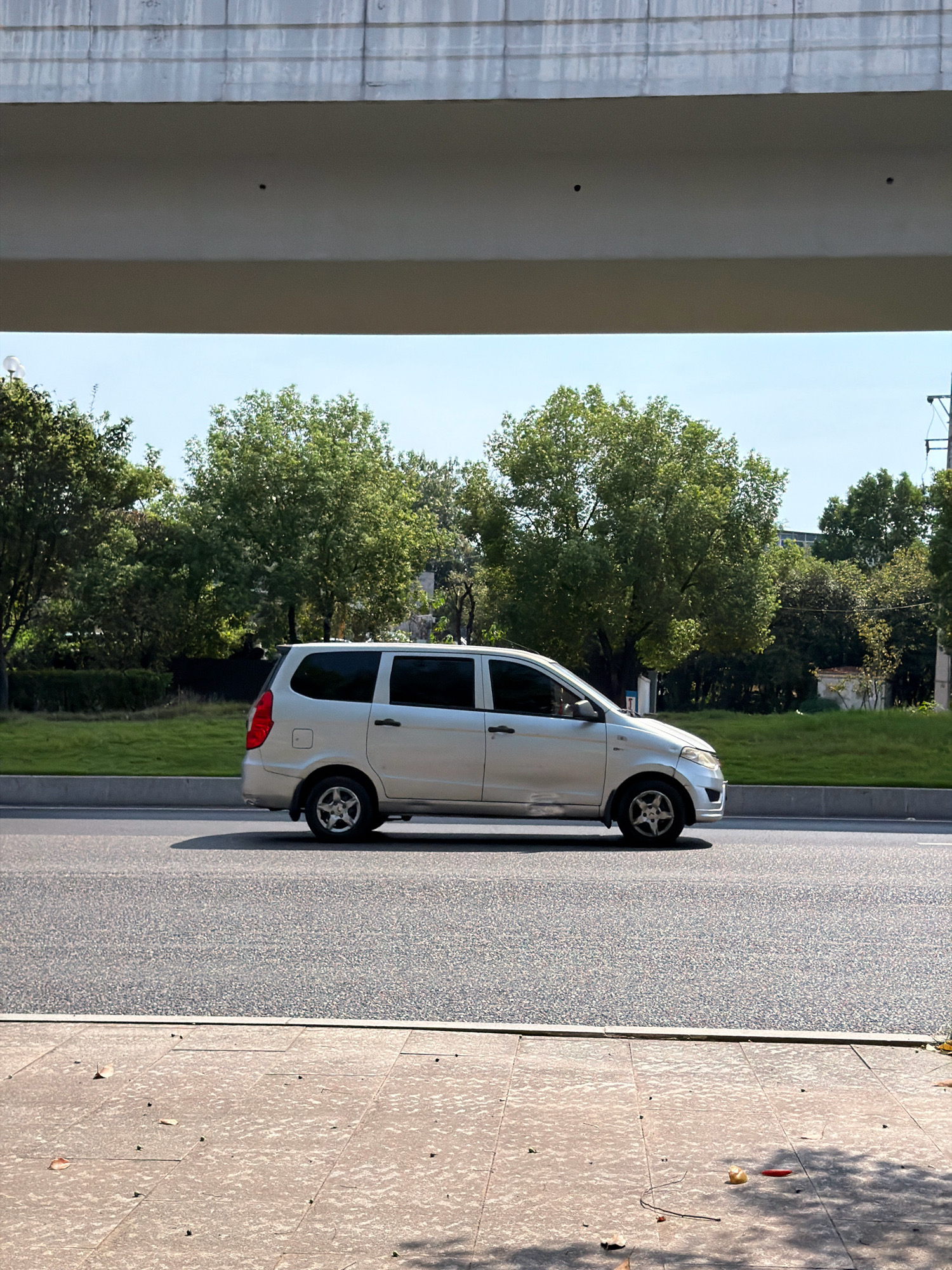 The image depicts a silver minivan driving on a road in Jiangjin, China. The minivan is traveling from left to right, positioned in the center lane of a multi-lane road. The road surface is asphalt. To the left of the road, there is a grassy median with trees. To the right of the road, more trees and some buildings are visible in the background. A concrete overpass or bridge structure extends across the top of the image. The sky is clear and blue. The foreground shows a paved sidewalk made of square tiles with some fallen leaves scattered on it. The lighting suggests it is daytime, likely with bright sunlight given the shadows.