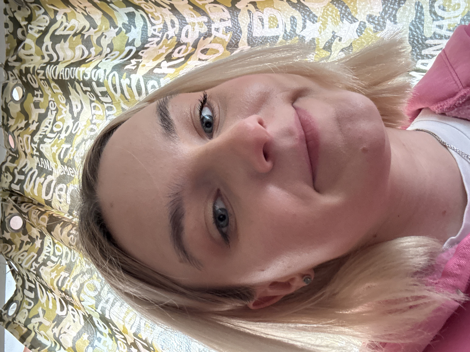 The image shows a close-up view of a person with light blonde hair and blue eyes. The person is positioned at an angle, with their face slightly tilted. A patterned curtain hangs in the background. The person is wearing a pink top and a white undershirt with a silver chain. The location and specific city are unknown.
