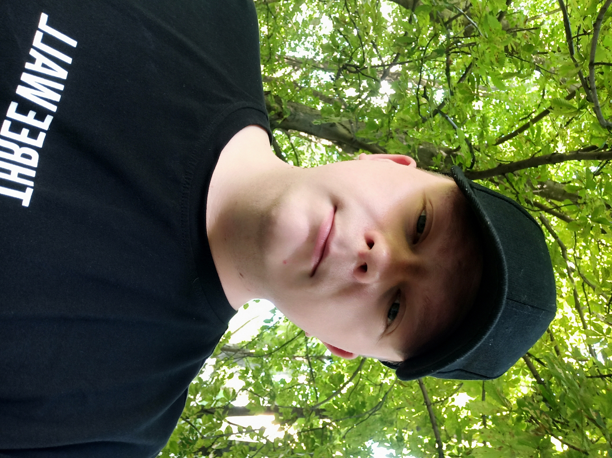 A young person wearing a black baseball cap and a black t-shirt stands outdoors. The t-shirt has white text on it that appears to be reversed. The person's face is in focus, and they are looking directly at the camera. Behind them, lush green leaves and tree branches fill the frame, suggesting they are in a wooded or park-like area. The sunlight filters through the leaves, creating a dappled effect. The setting appears to be outdoors during daylight.